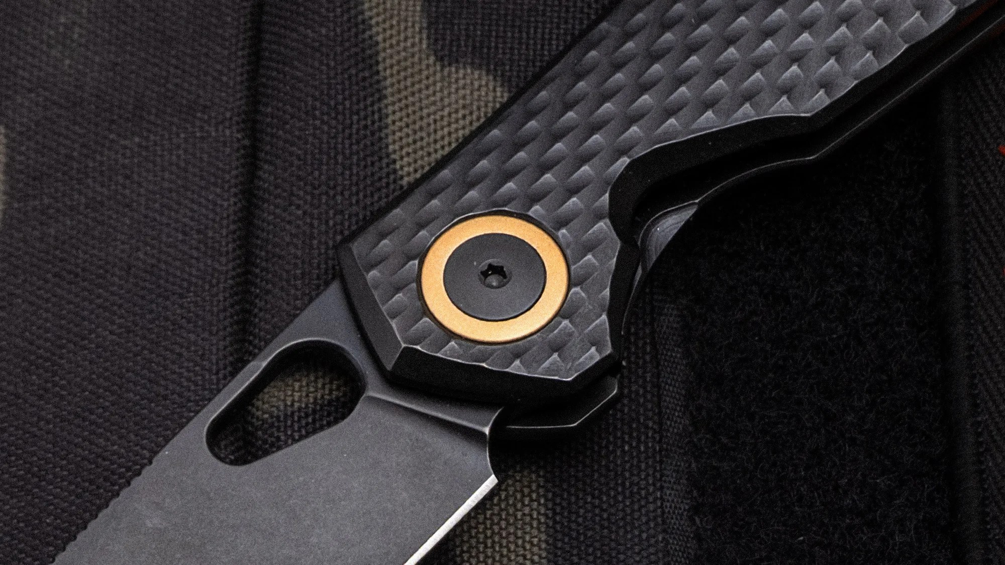 Close-up of a black folding knife with a textured carbon fiber handle and a gold-accented pivot screw.