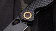 Close-up of a black folding knife with a textured carbon fiber handle and a gold-accented pivot screw.