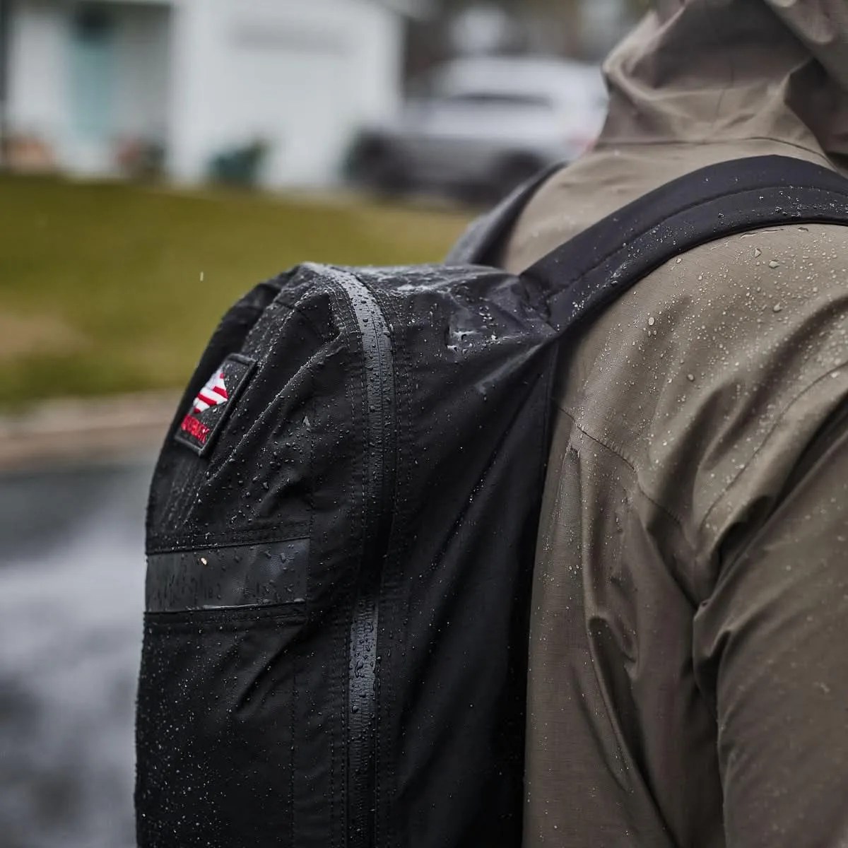 Black waterproof backpack with water droplets worn over a brown rain jacket.