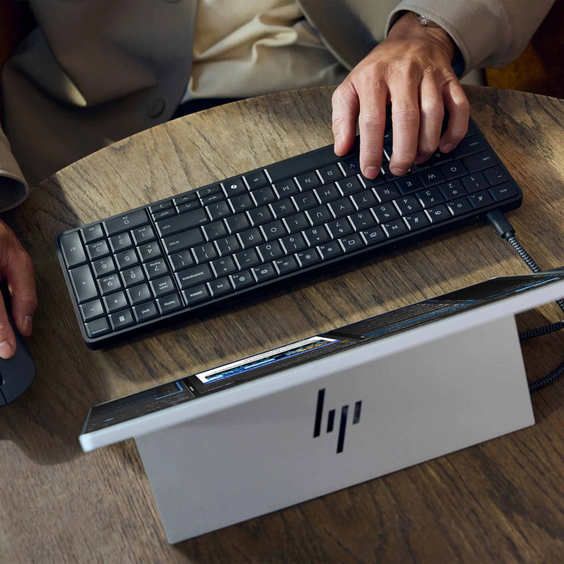 Person typing on a black compact keyboard connected to a silver HP laptop on a wooden table.
