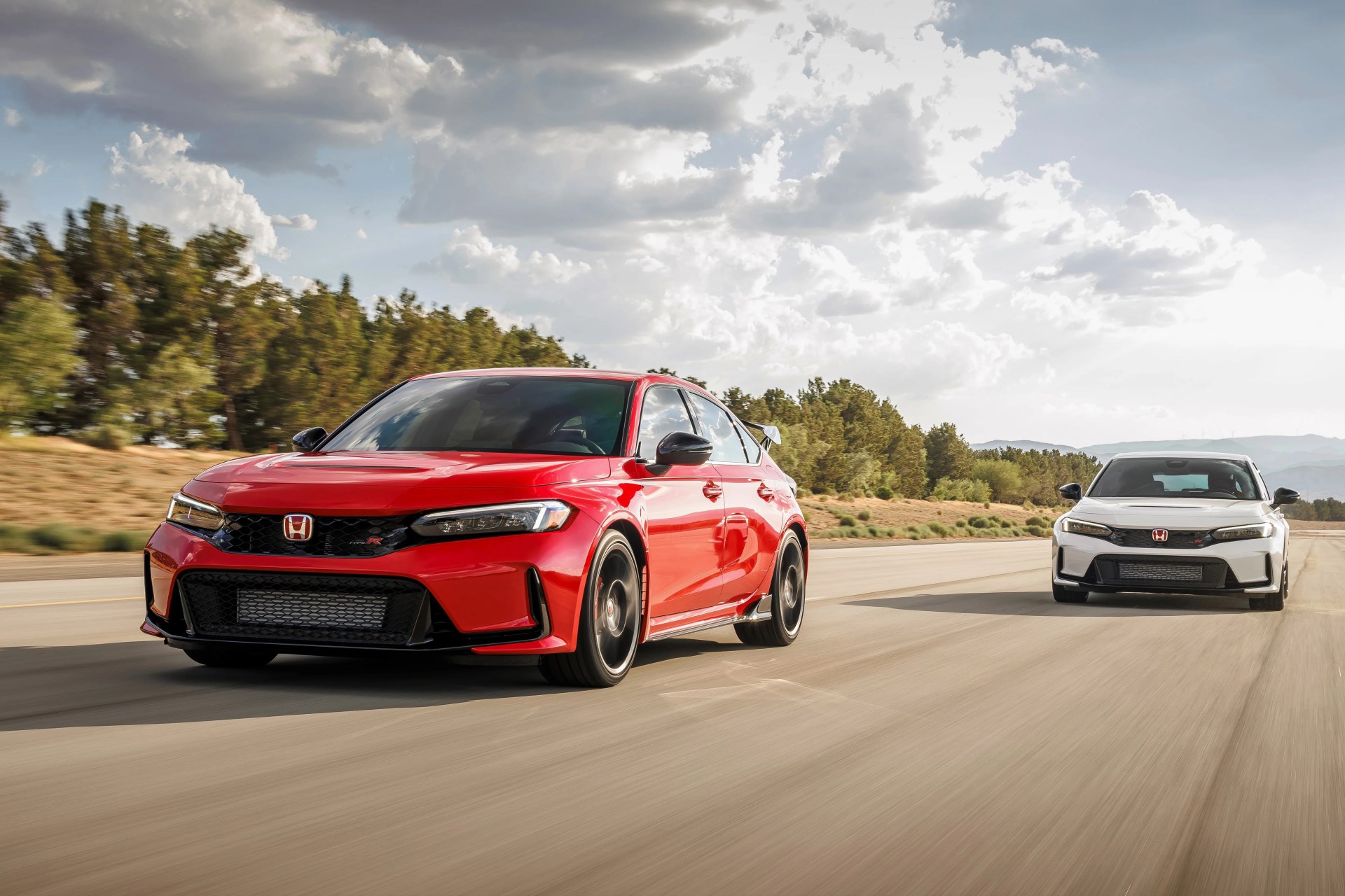 Red and white Honda Civic Type R cars driving on a road with trees and cloudy sky in the background.