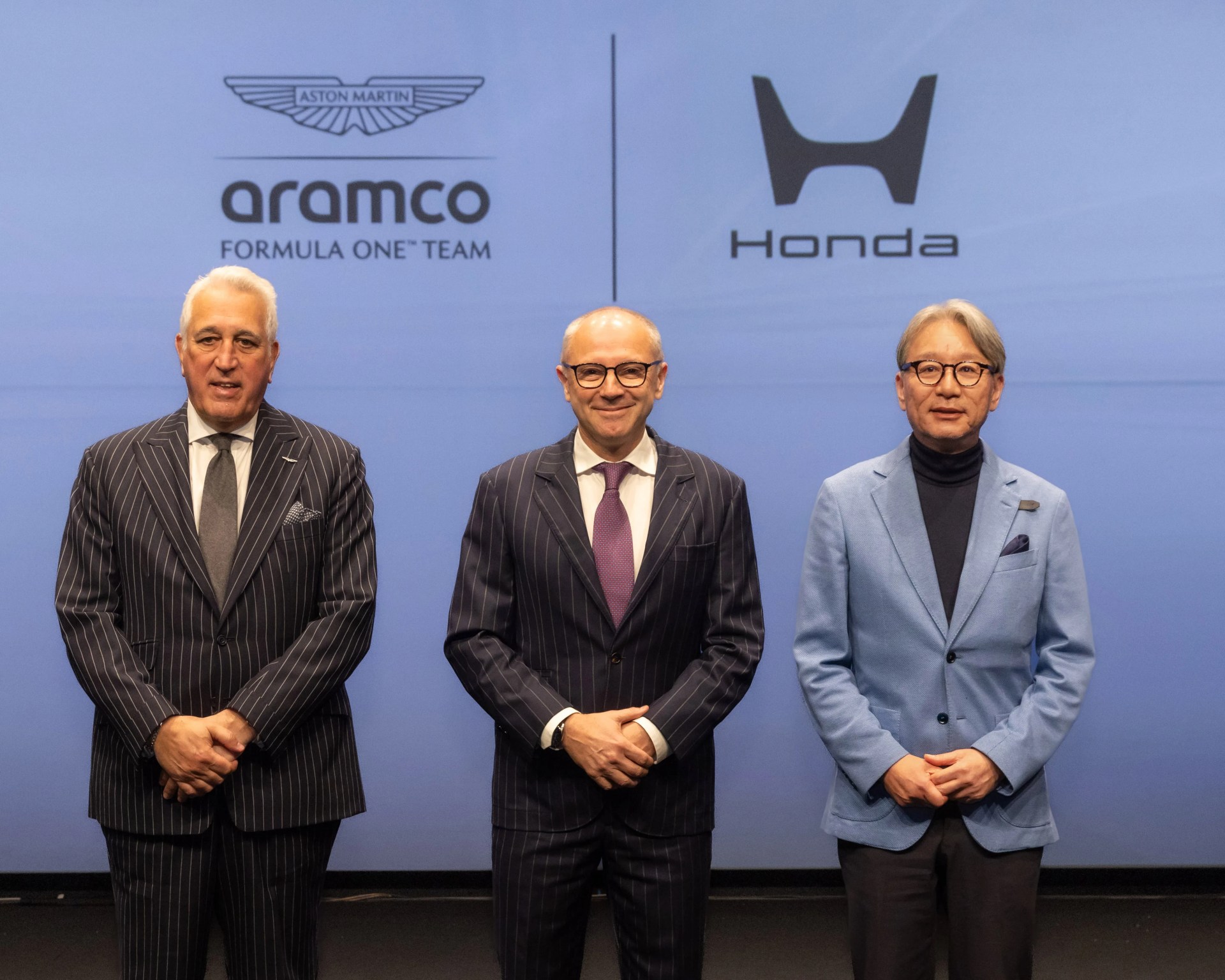 Three men in suits standing in front of a blue backdrop with Aston Martin Aramco Formula One Team and Honda logos.