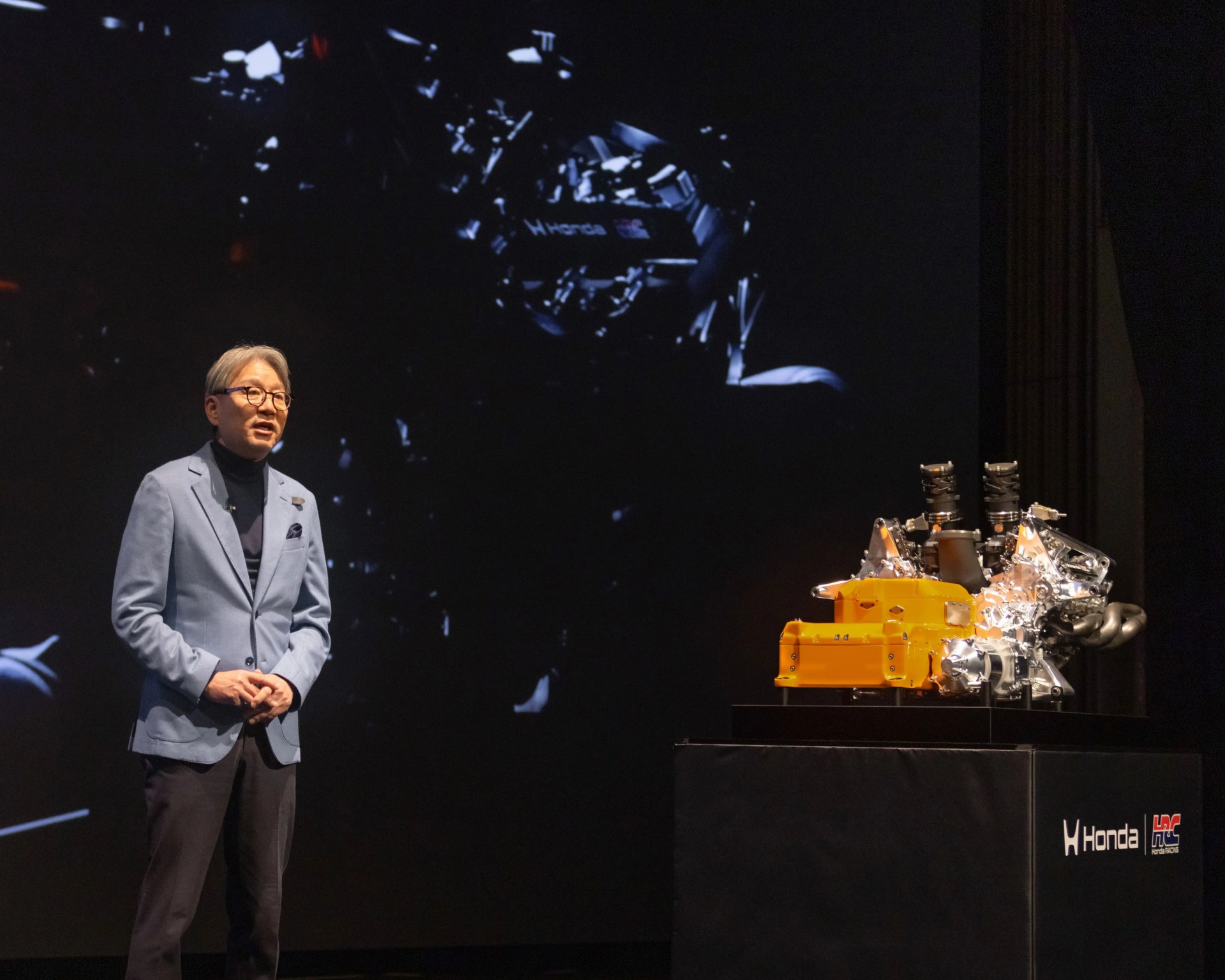 Man in blue blazer standing next to a Honda engine model on a black pedestal with Honda and HRC logos.