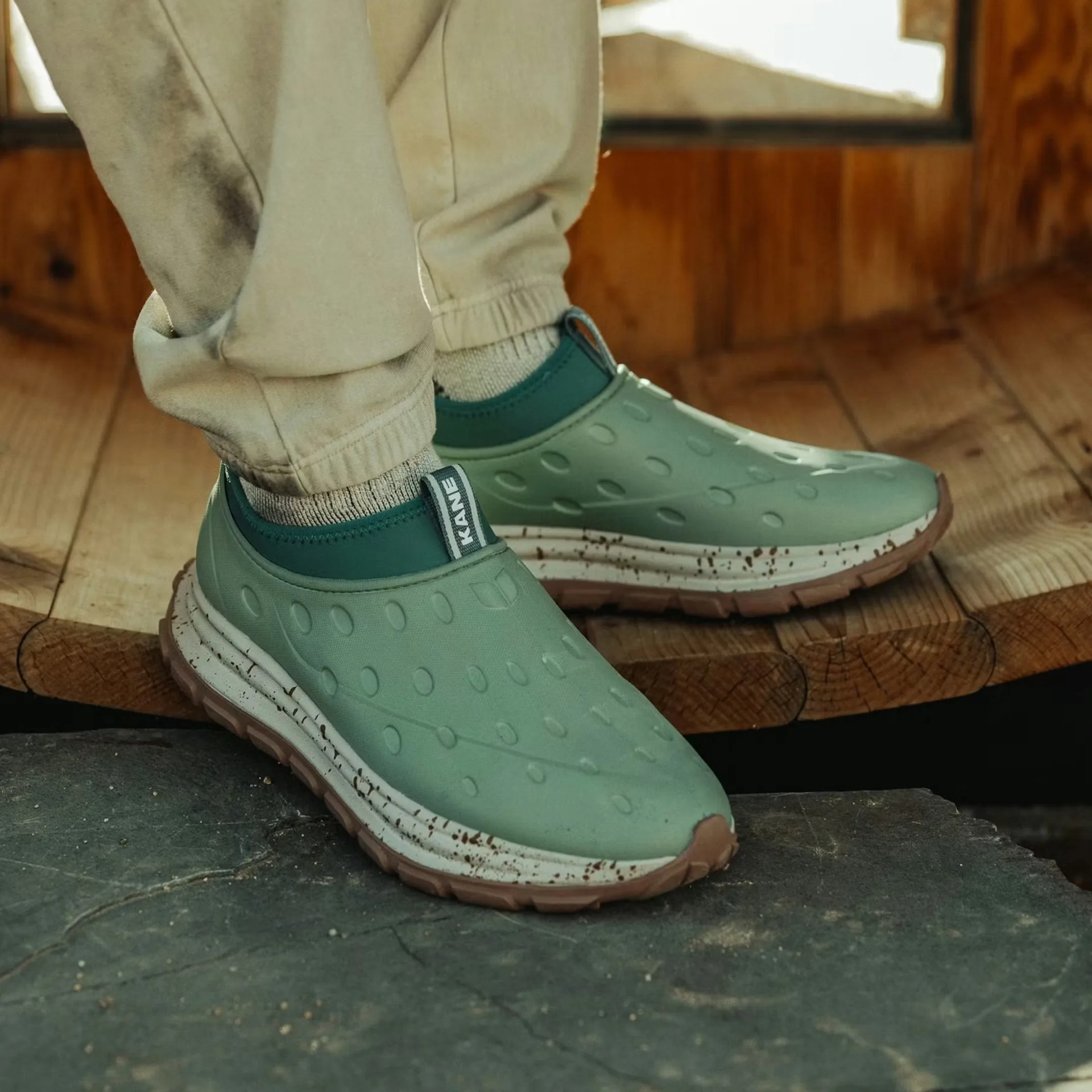 Green slip-on sneakers with textured upper and white midsoles worn with beige pants on wooden surface.