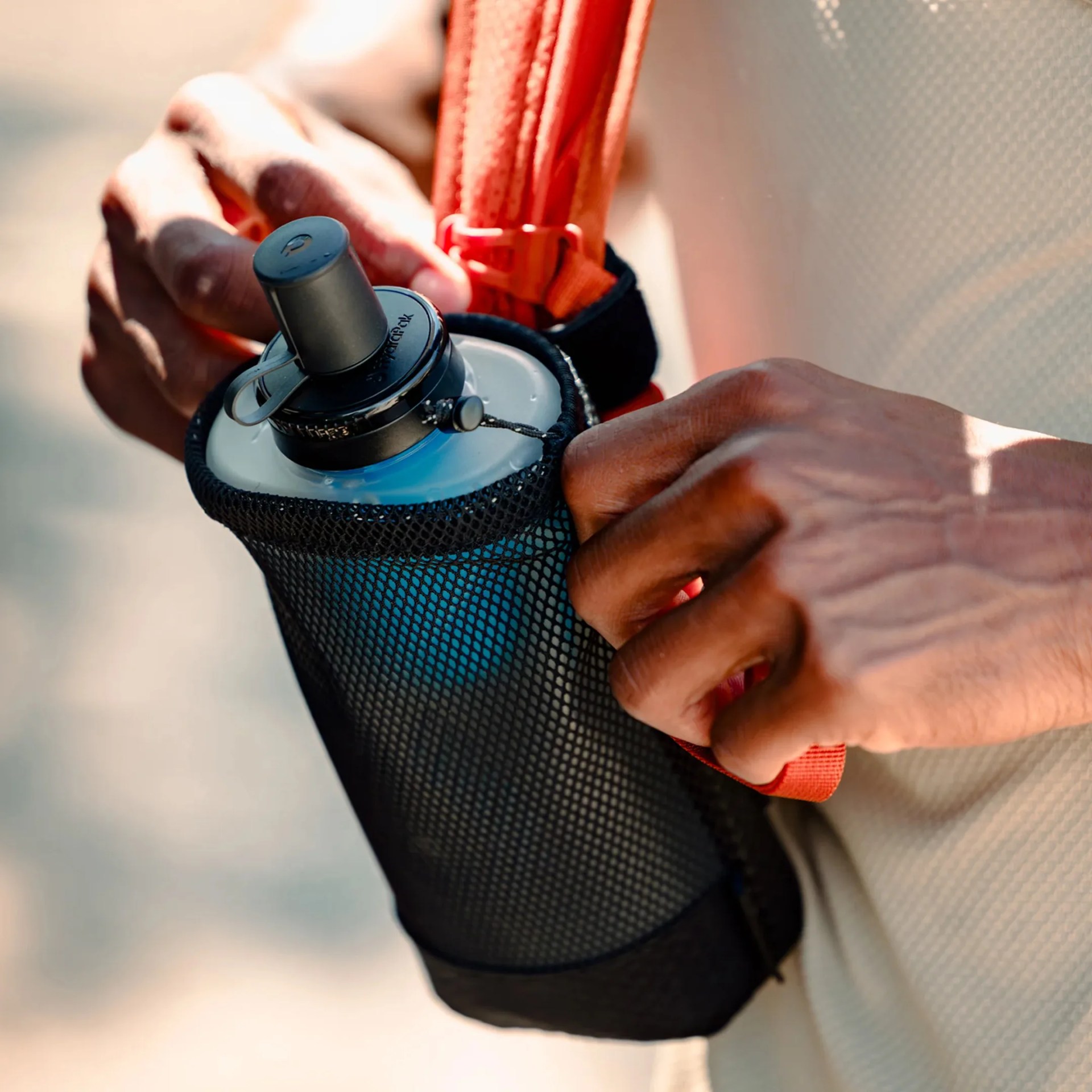 Black mesh water bottle holder with a gray bottle inside, held by a person wearing a light-colored shirt.