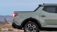 Rear side view of a light gray Hyundai pickup truck with black trim and alloy wheel on a gravel road with desert landscape in the background.