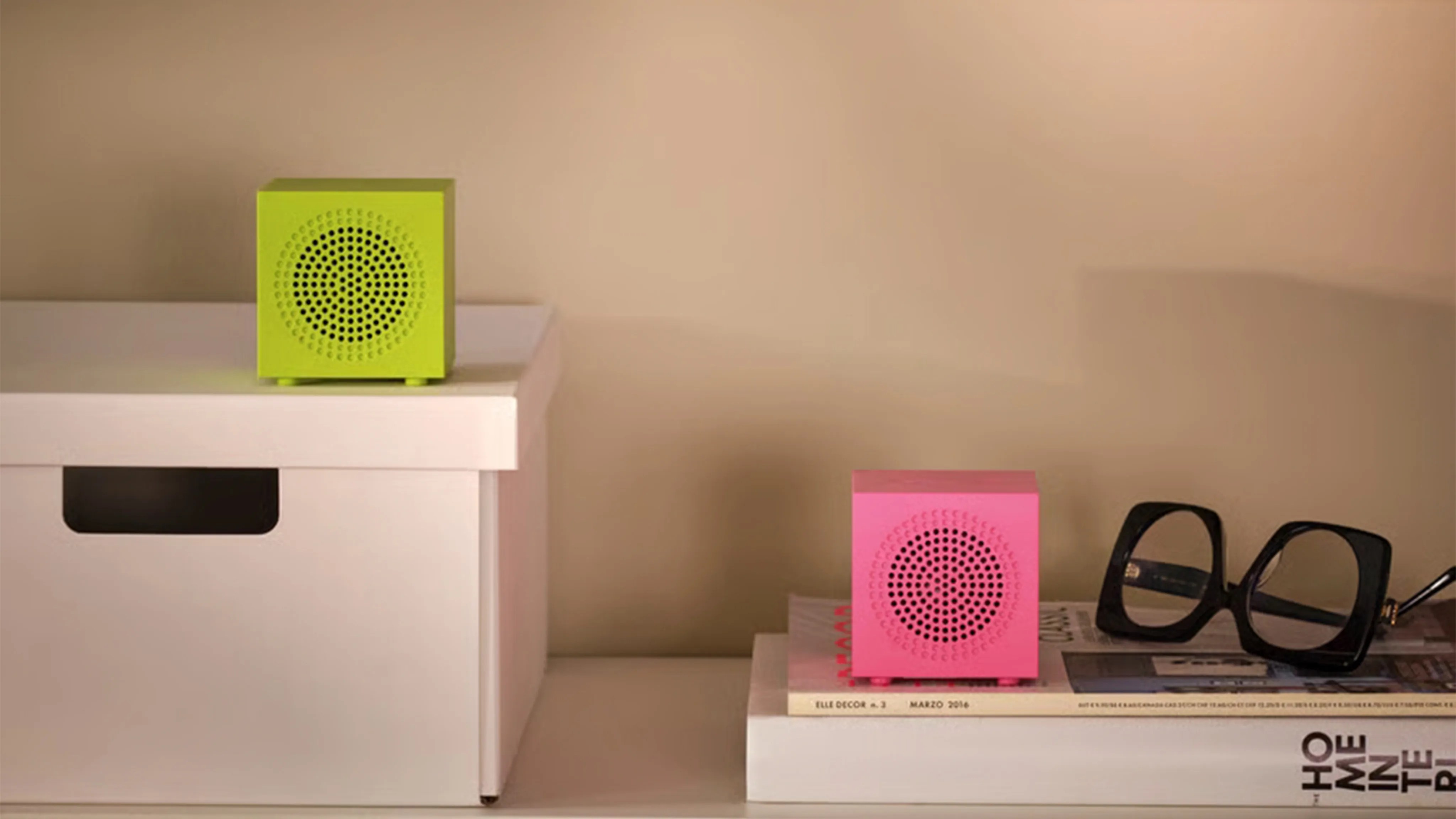 Two small cube-shaped speakers, one lime green on a white box and one pink on stacked books next to black square-framed glasses.
