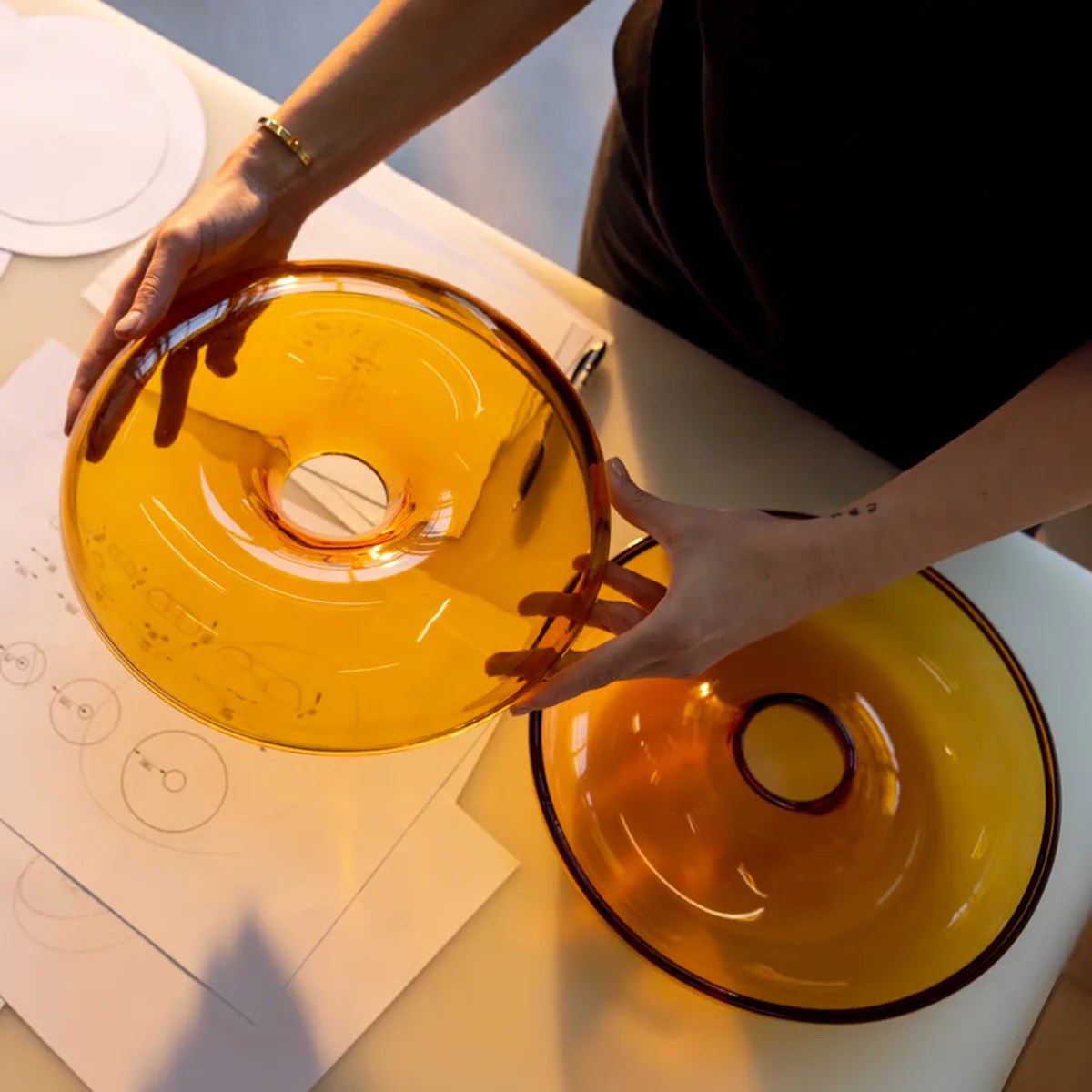Two amber-colored glass bowls with a central hole, one held in hands and the other on a table with design sketches.