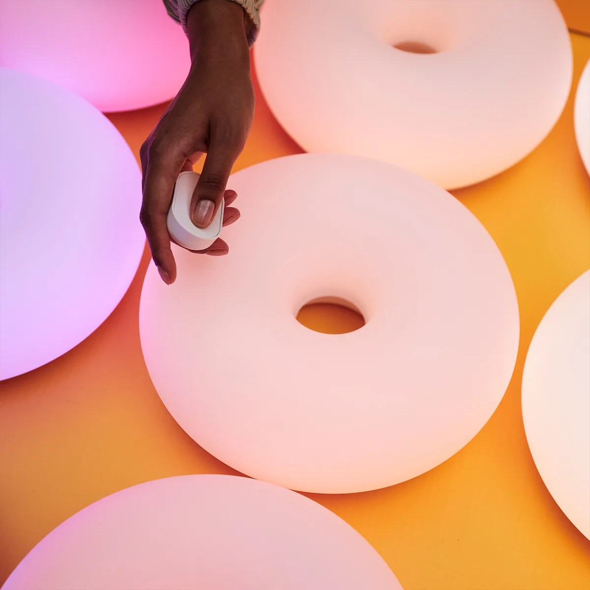 Hand holding a small white remote over round, donut-shaped glowing lights on an orange surface.