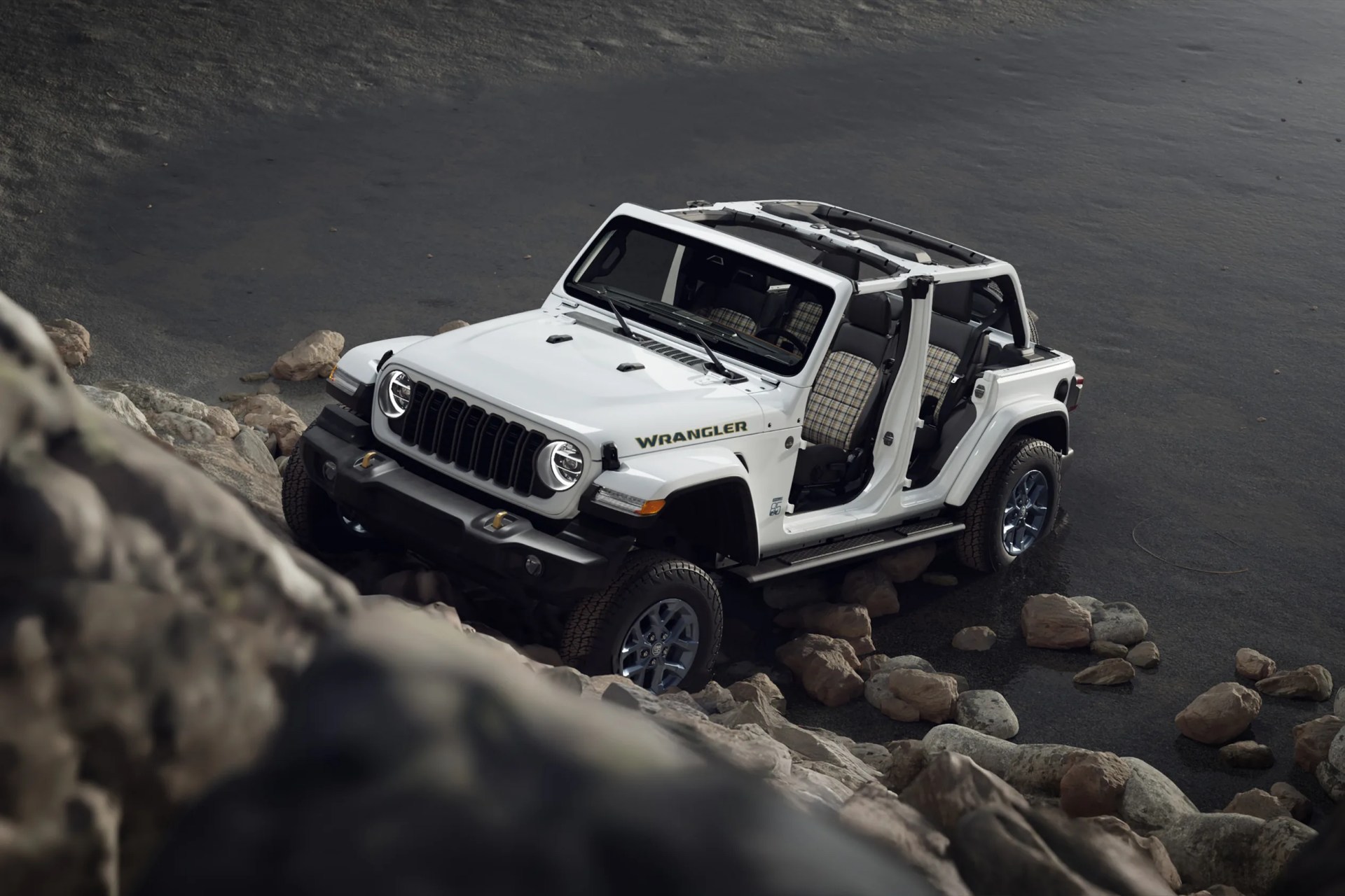 White Jeep Wrangler with open doors and plaid seats climbing over rocks on a dark gravel surface.