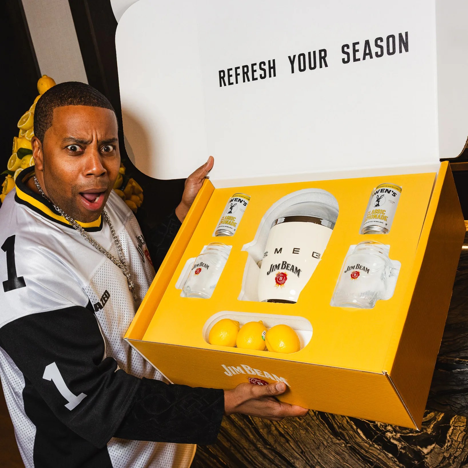 Person holding an open yellow Jim Beam gift box containing a white ice bucket, two glass mugs, two cans of lemonade, and three lemons.
