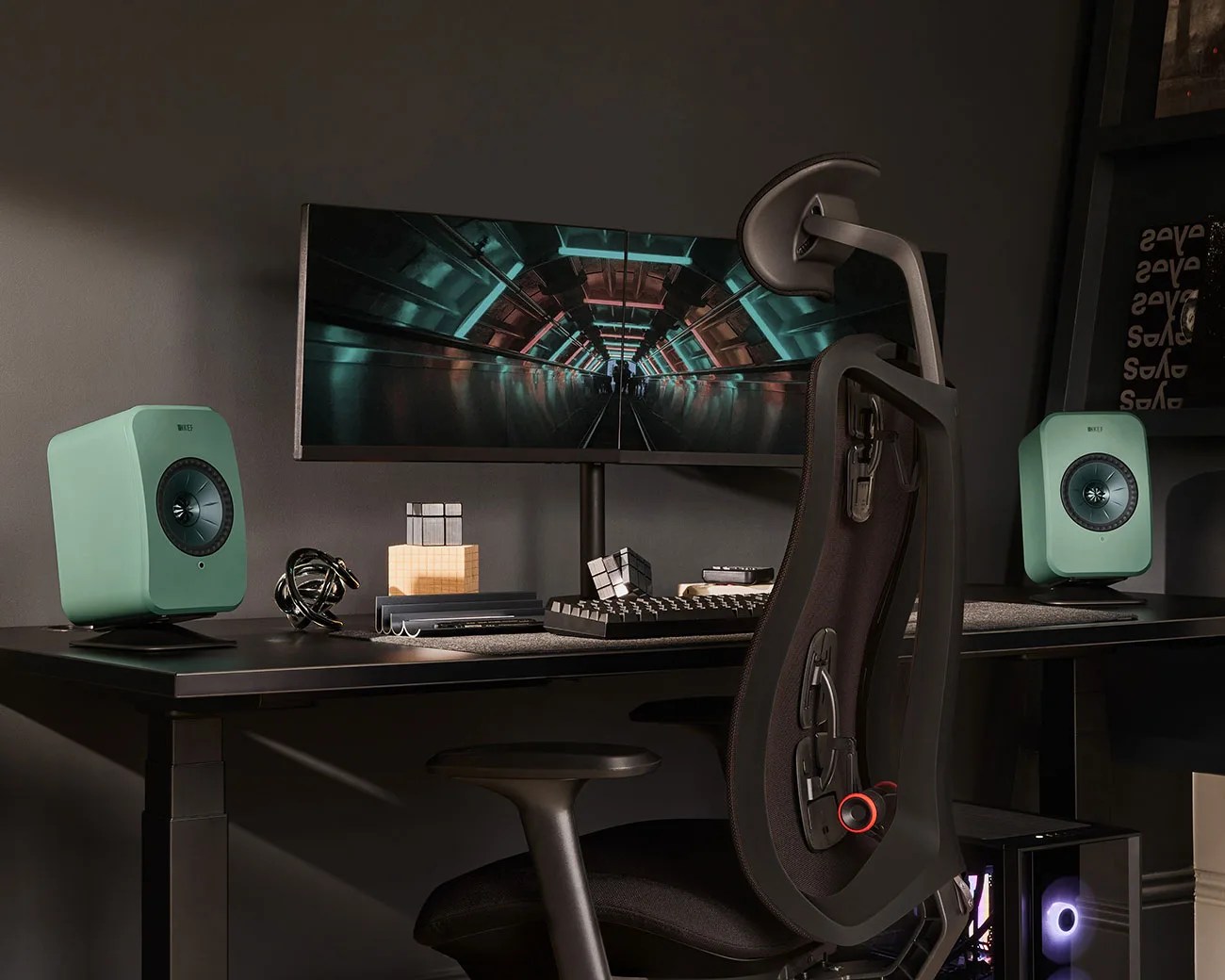 Black ergonomic office chair in front of a black desk with a curved ultrawide monitor and two green KEF speakers.