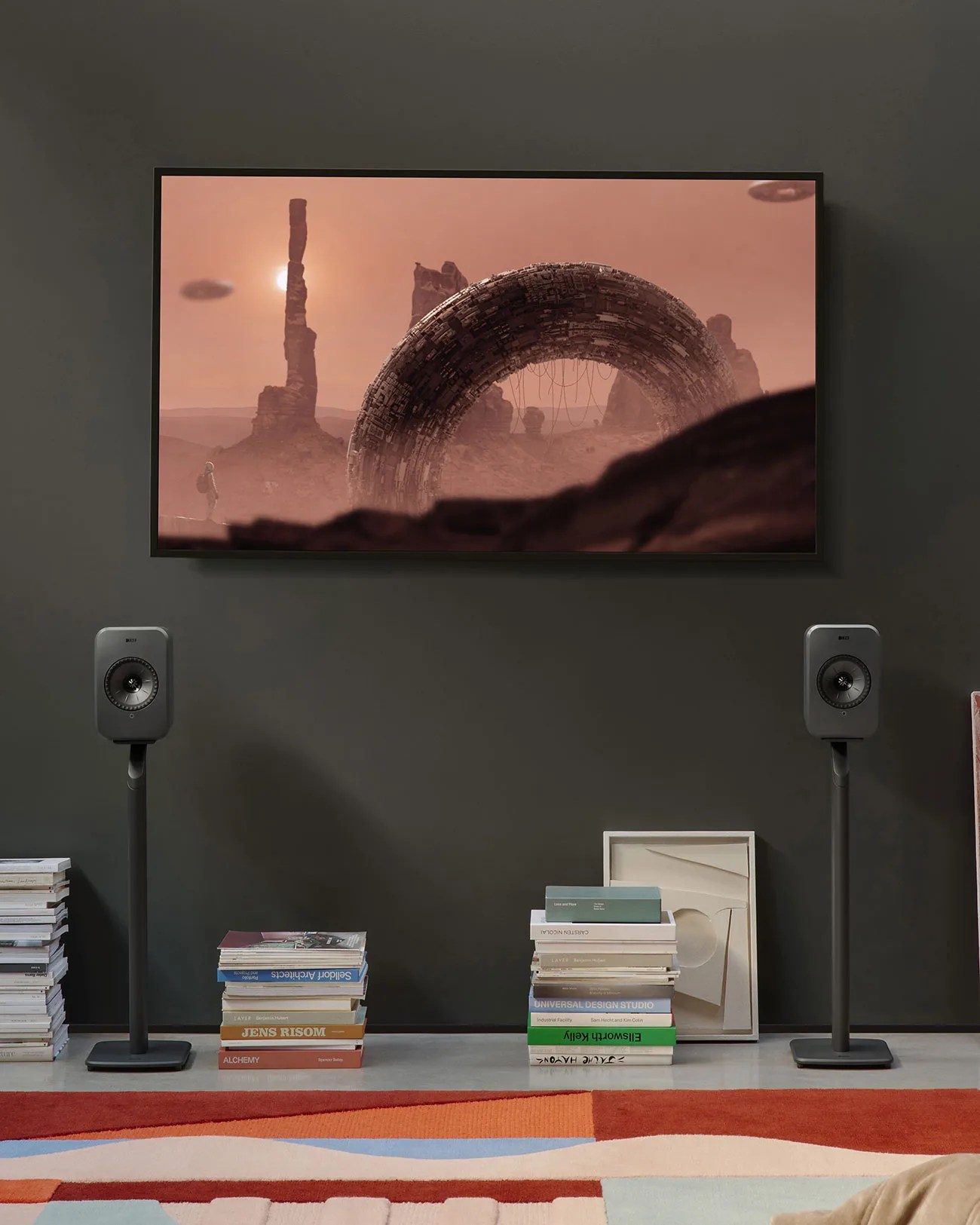 Two black KEF speakers on stands flanking a wall-mounted TV displaying a sci-fi scene with a large circular structure and rocky landscape.