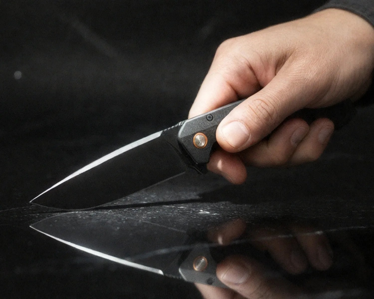 Hand holding a black folding knife with a sharp blade reflected on a dark surface.