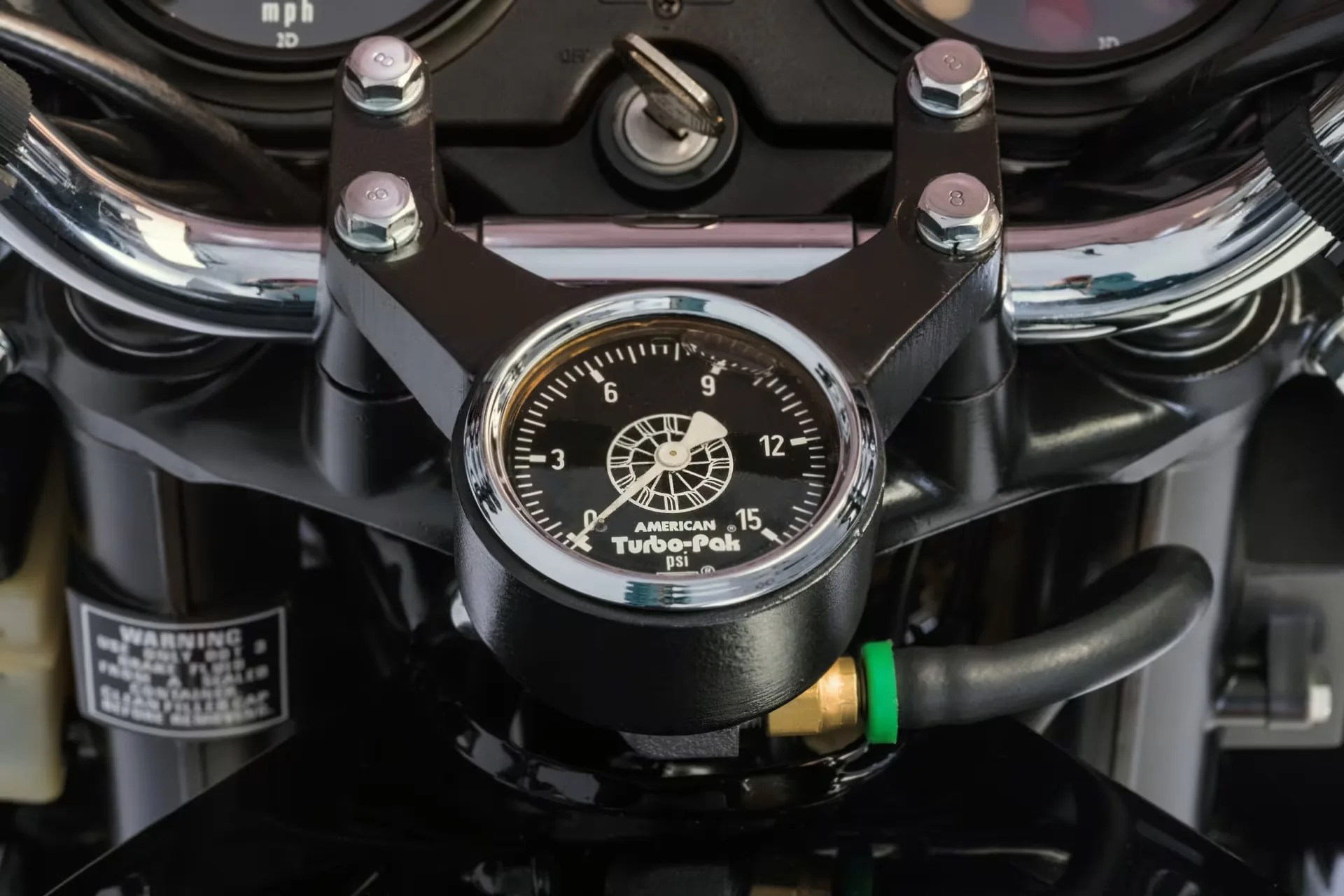 Black American Turbo-Pak psi gauge mounted on a motorcycle handlebar with chrome bolts and a black hose attached.