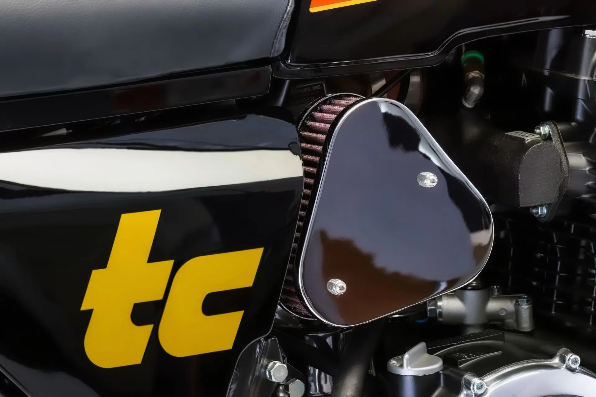 Close-up of a black motorcycle air filter cover with a yellow "tc" logo on the adjacent black body panel.