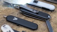 Black multi-tool with scissors and knife blades extended, lying on a wooden surface.