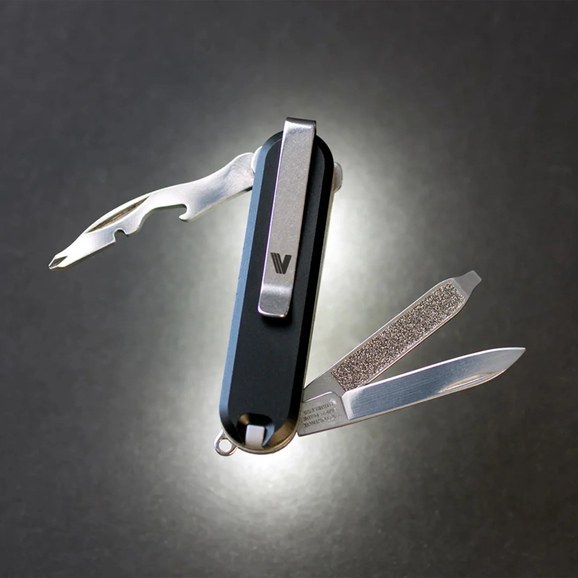 Black multi-tool with metal clip, opened to show a bottle opener, nail file, and small knife blade.