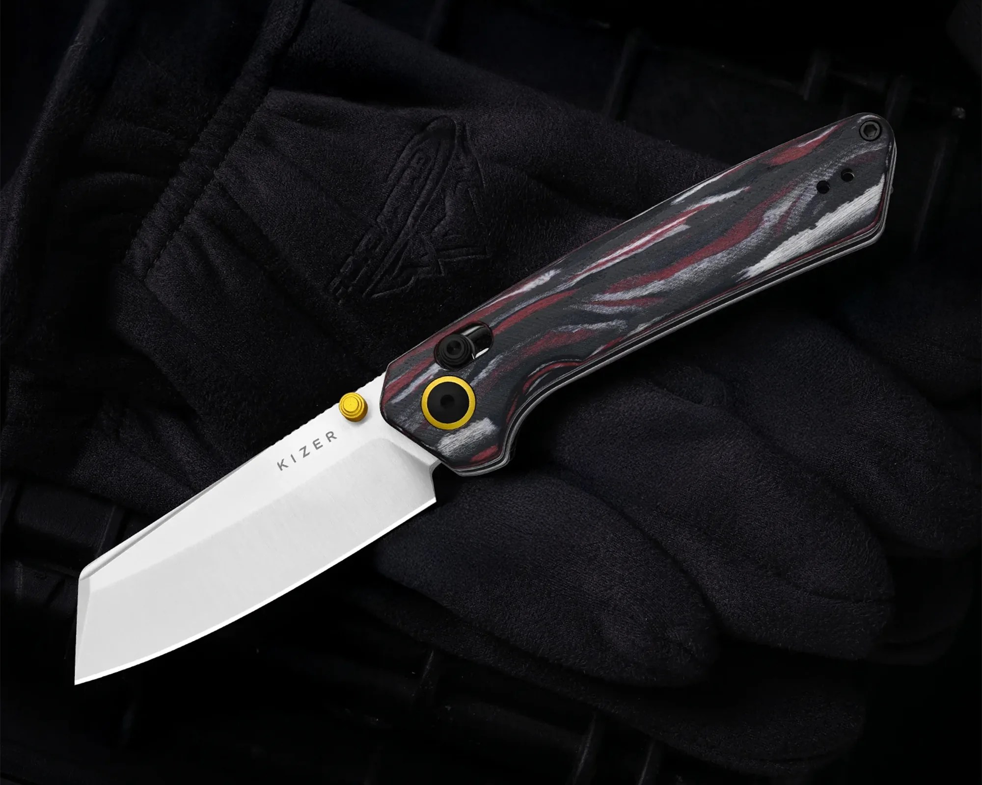 Folding knife with a silver blade labeled "KIZER" and a black, red, and gray patterned handle resting on black fabric.