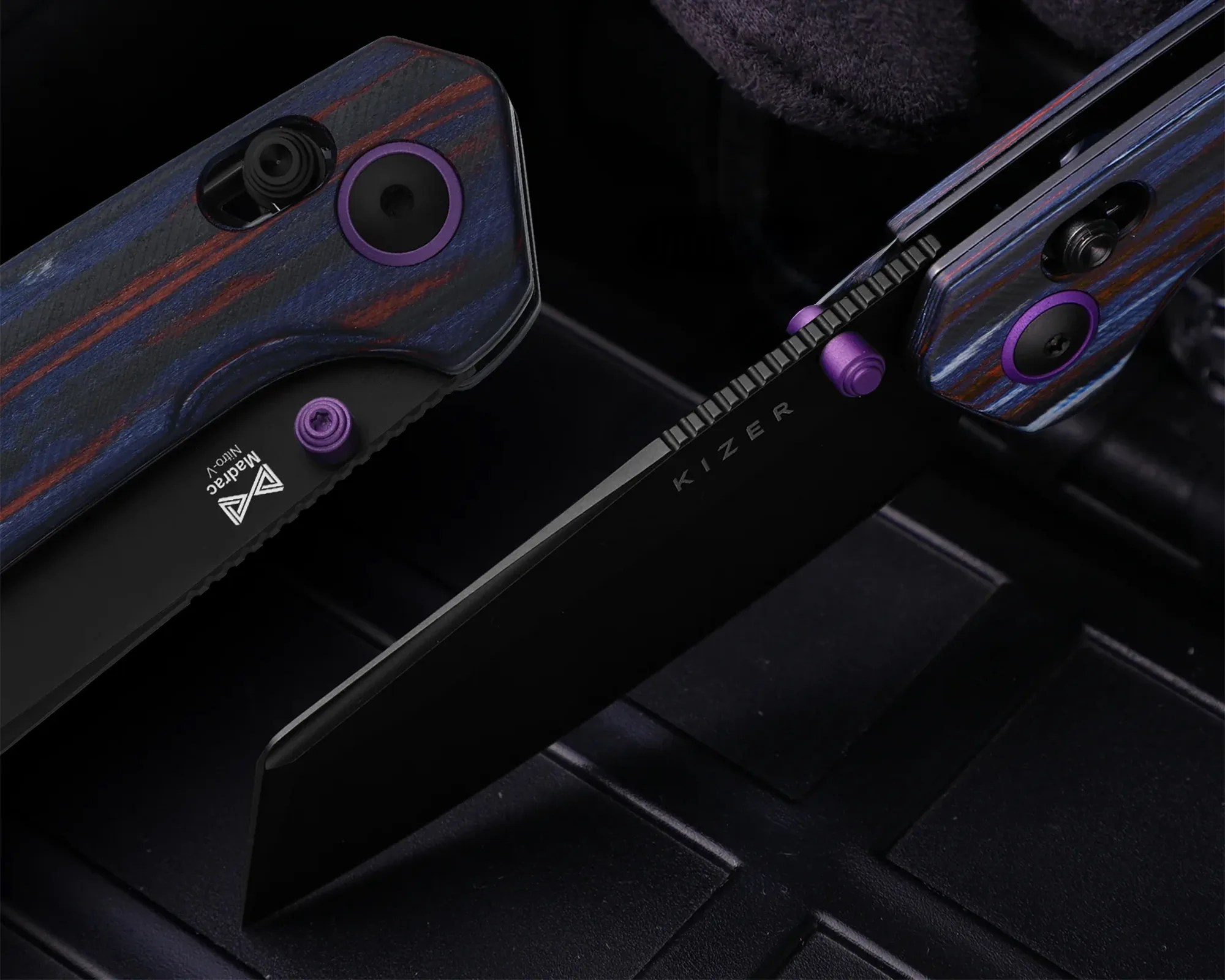 Two folding knives with black blades, purple thumb studs, and blue and red layered handles on a black surface.
