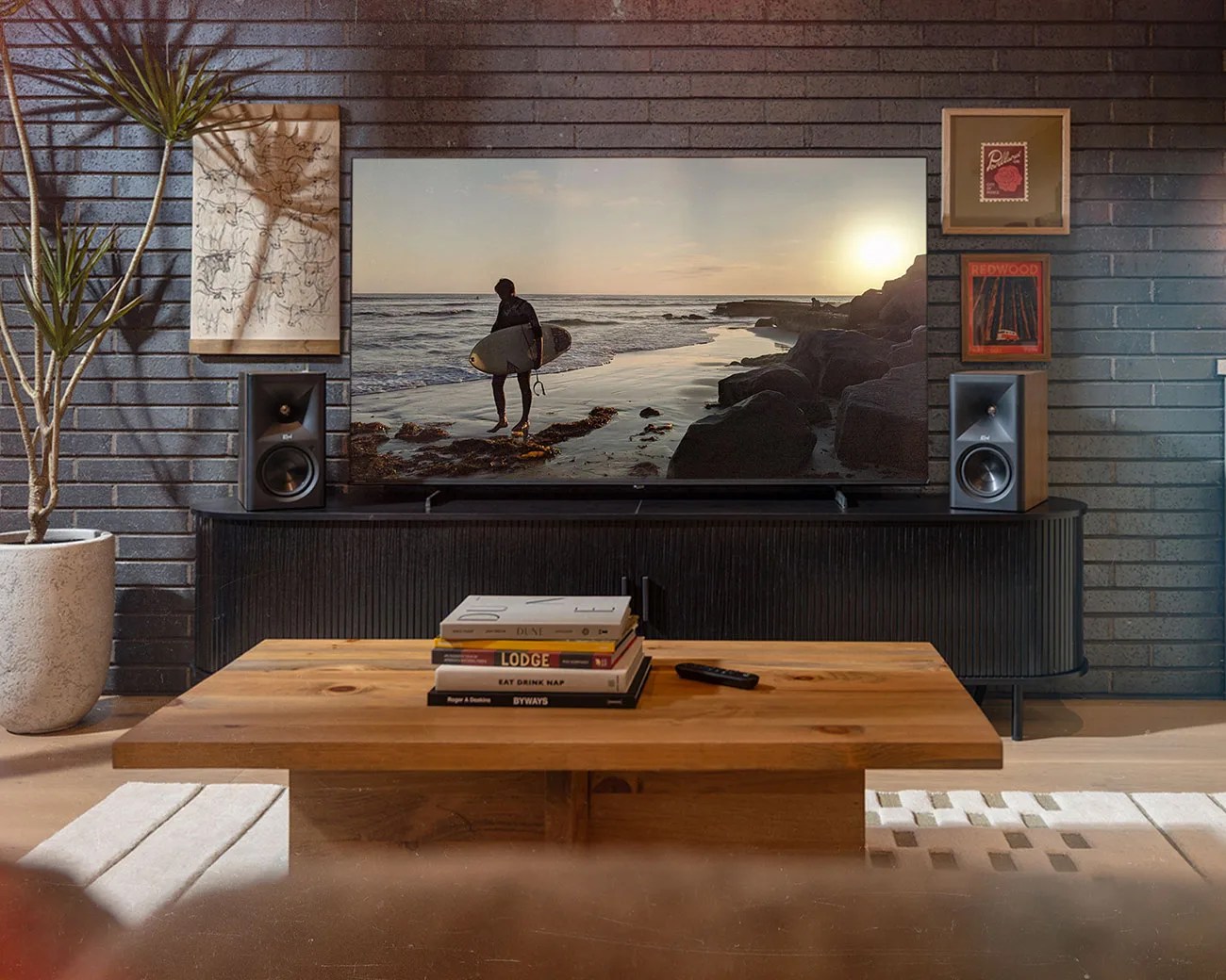 Living room with a large flat-screen TV on a black media console, flanked by two speakers, a wooden coffee table with stacked books, and a potted plant.