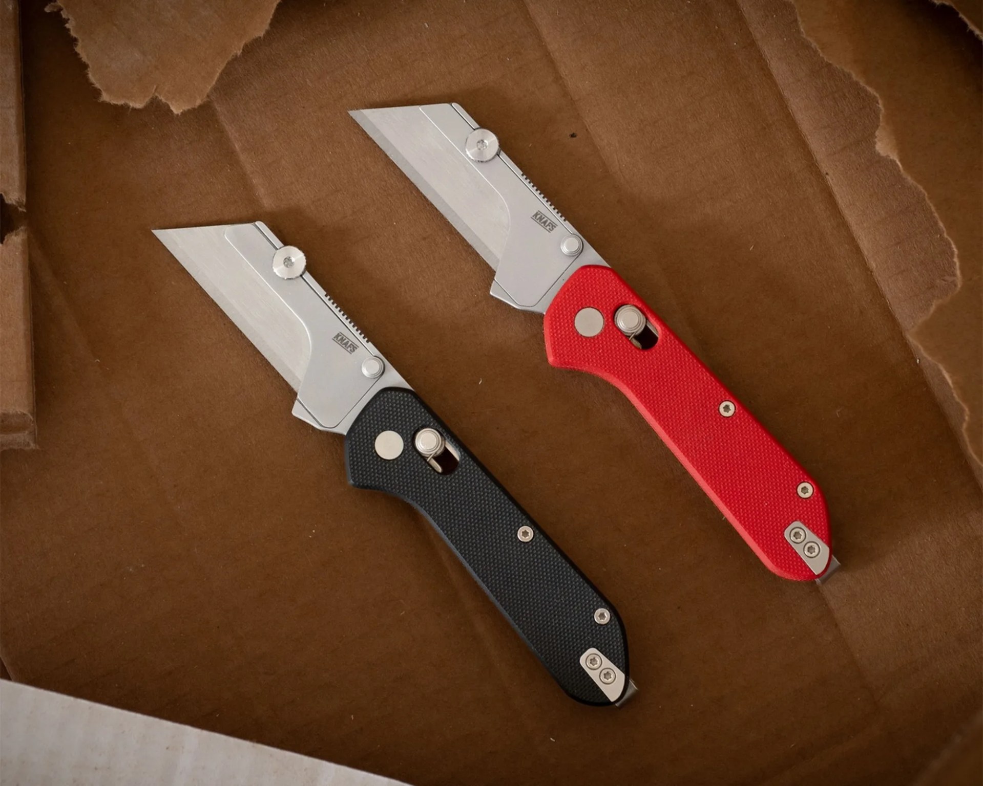 Two utility knives with silver blades, one with a black textured handle and one with a red textured handle, on a brown cardboard surface.