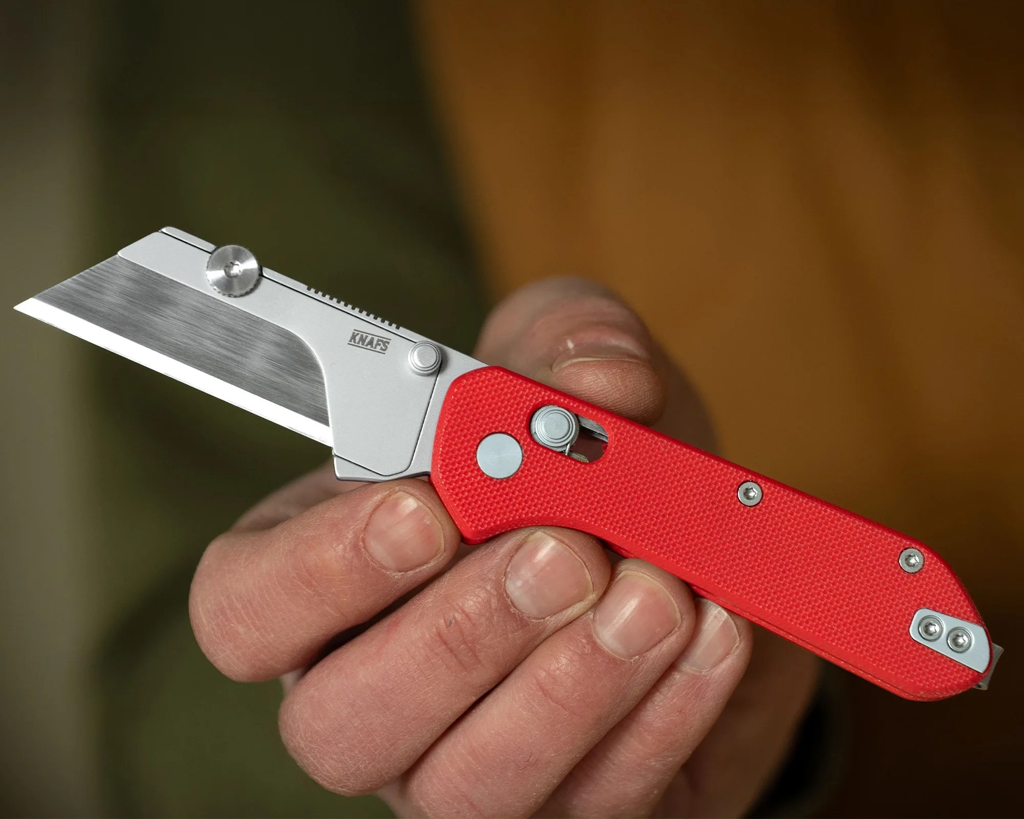 Hand holding a folding utility knife with a red textured handle and a silver blade marked "KNAFS.
