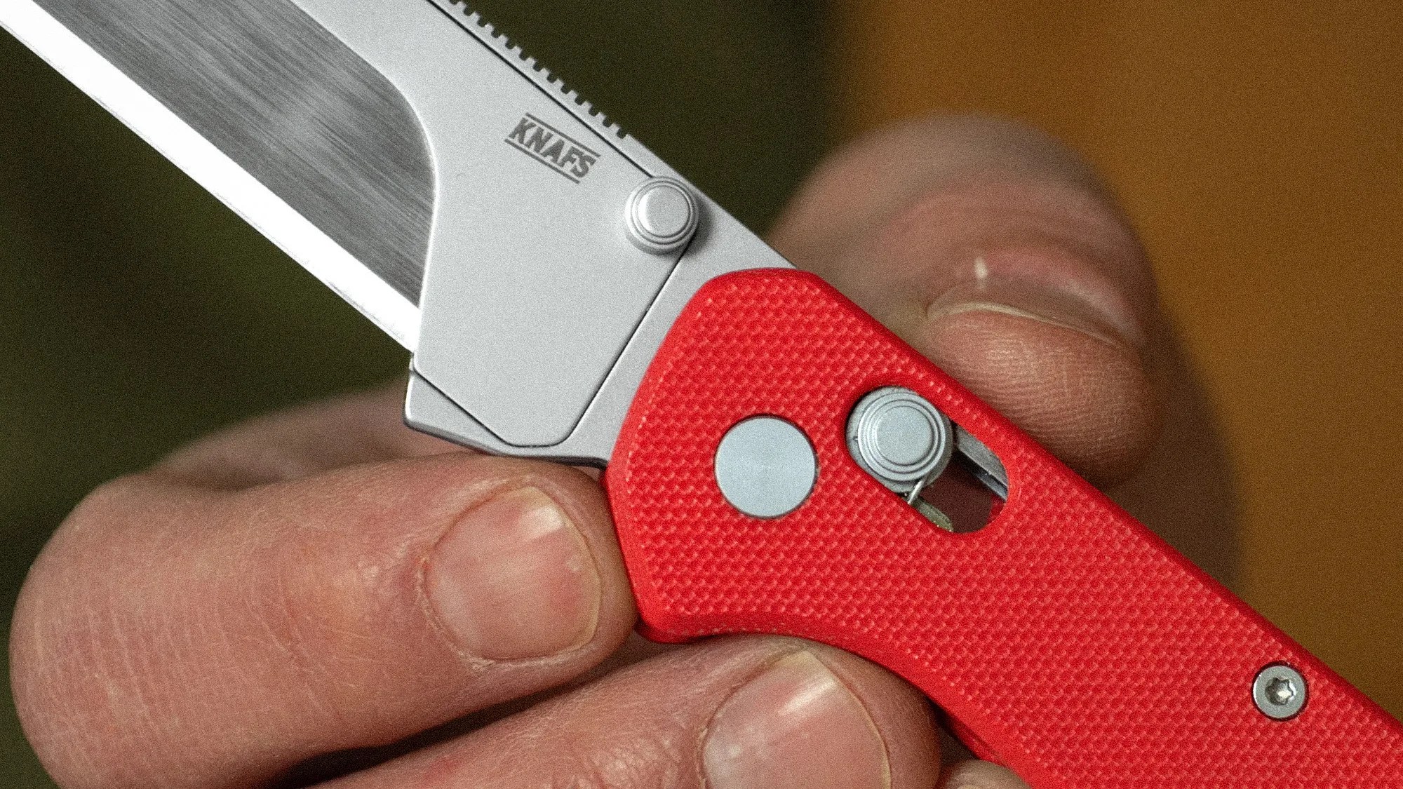 Close-up of a hand holding a folding knife with a red textured handle and a partially visible silver blade marked "KNAFS.