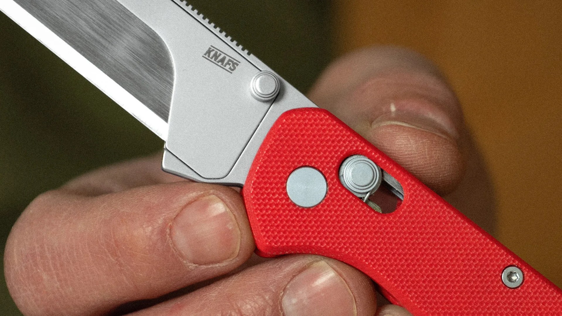 Close-up of a hand holding a folding knife with a red textured handle and a partially visible silver blade marked "KNAFS.