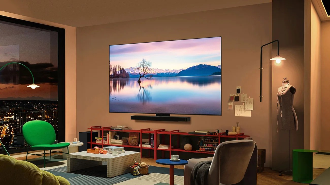 Living room with a large wall-mounted TV above a red shelving unit, green chair, gray armchair, and city view at dusk.