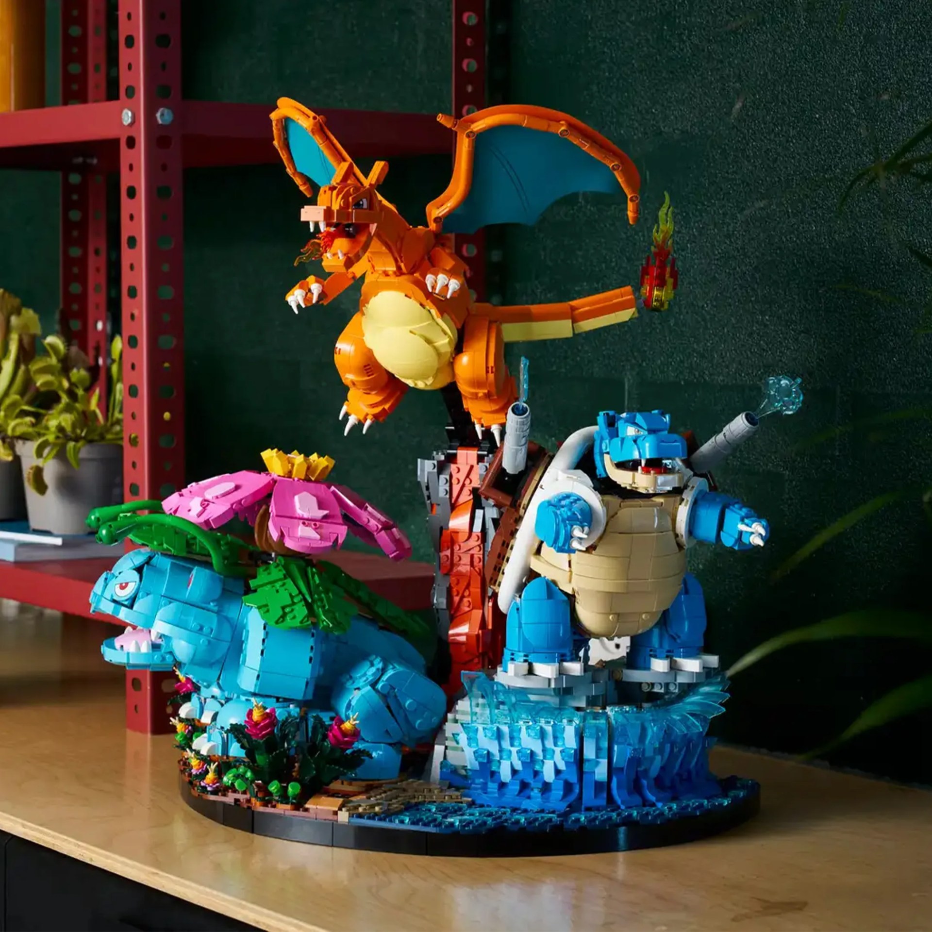 LEGO models of Pokémon Venusaur, Charizard, and Blastoise displayed on a wooden surface with a red metal shelf and plants in the background.