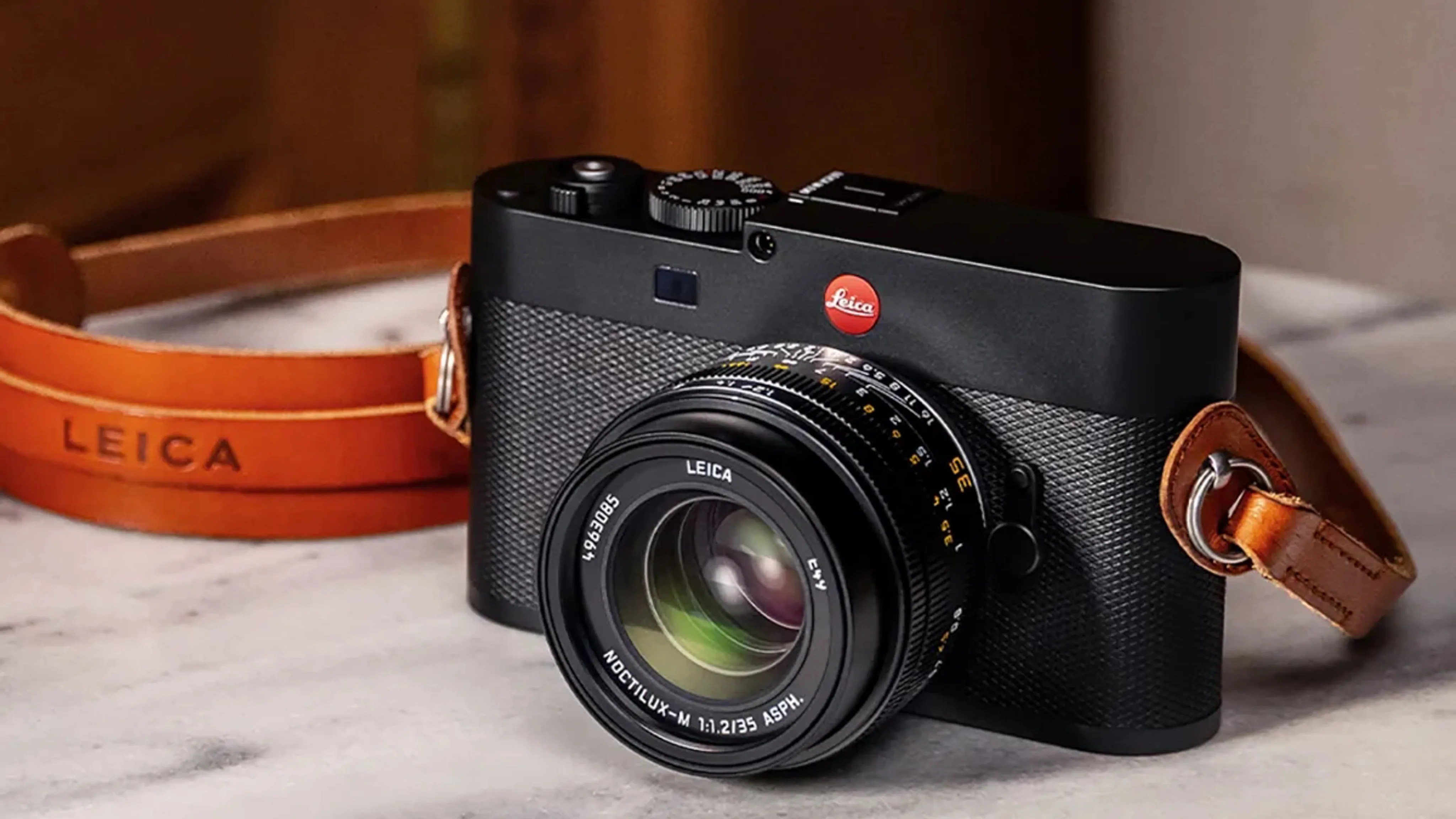 Black Leica camera with textured body and attached brown leather strap on a marble surface.