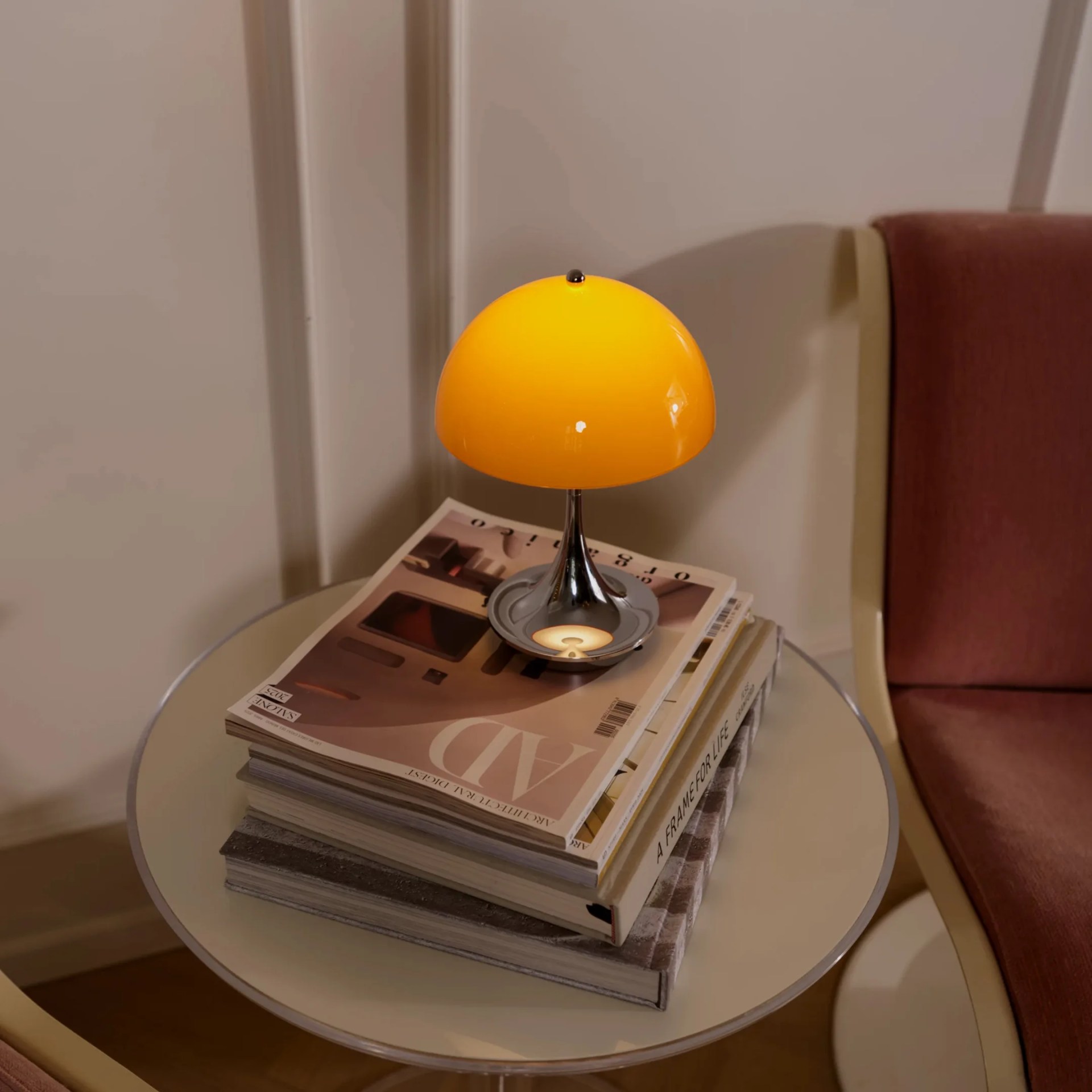 Small yellow dome-shaped table lamp with chrome base on a stack of magazines on a round glass table.