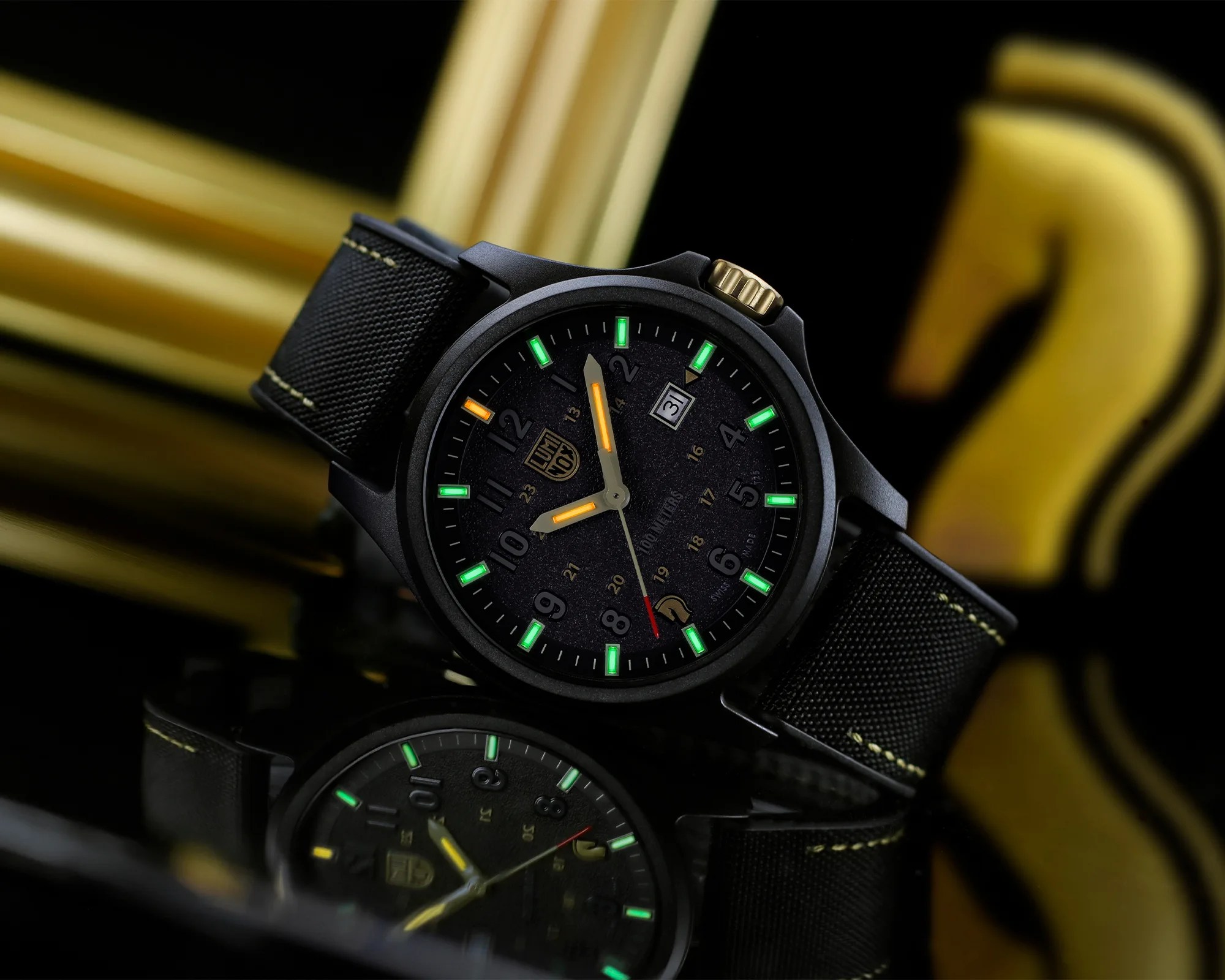 Black wristwatch with green and orange luminescent markers, black fabric strap, and gold crown on reflective surface.