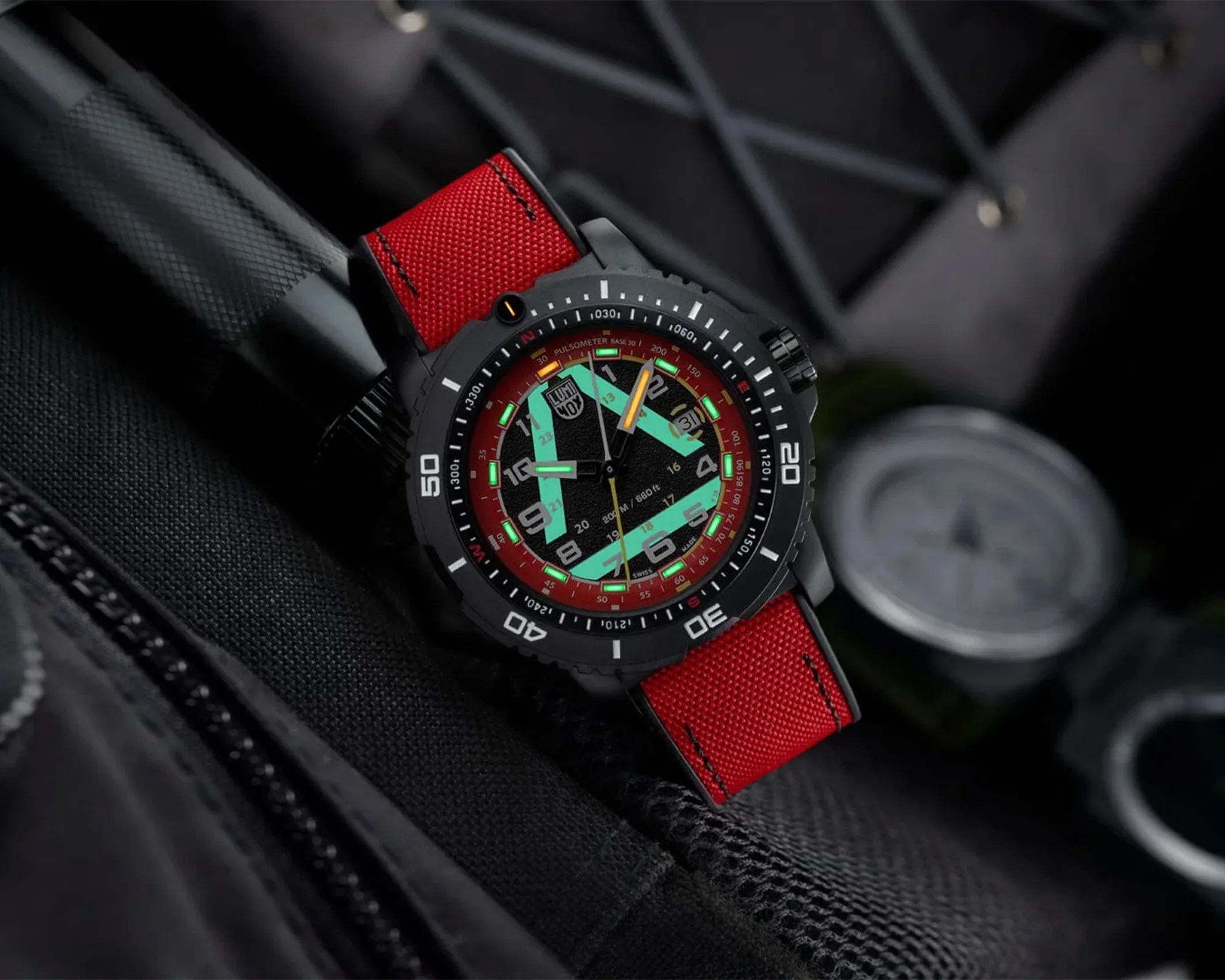 Black dive watch with red fabric strap and luminous green and yellow hands on a black textured surface.