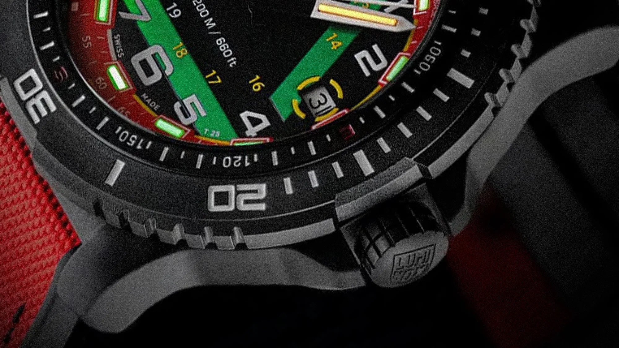 Close-up of a black watch bezel with white markings, a crown with "LUM-TEC" engraving, and a red fabric strap.
