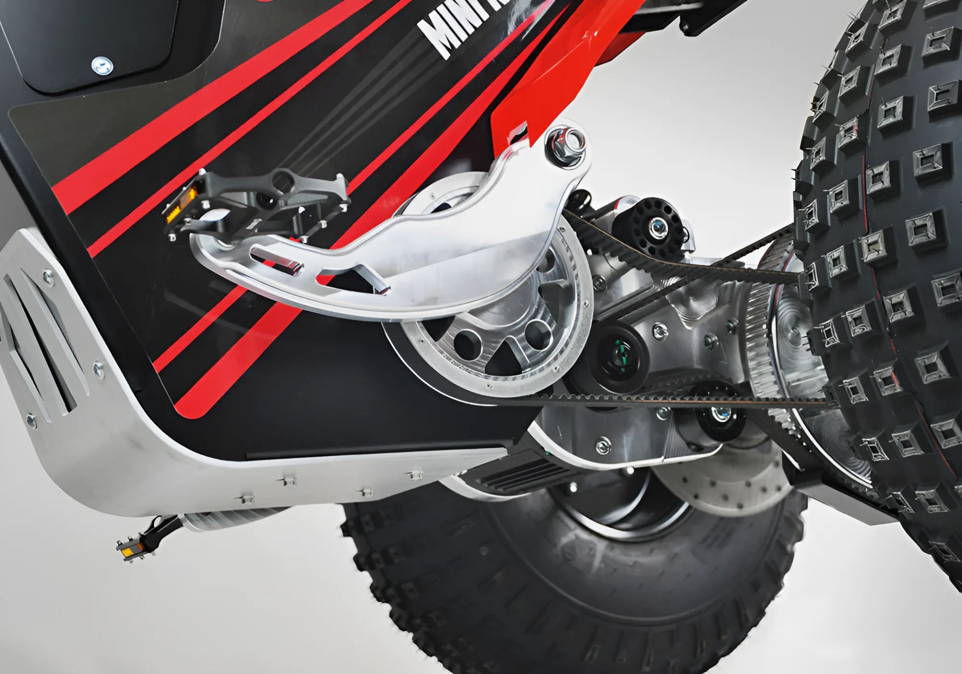 Close-up of a red and black electric dirt bike's pedal, chain drive, and knobby rear tire.