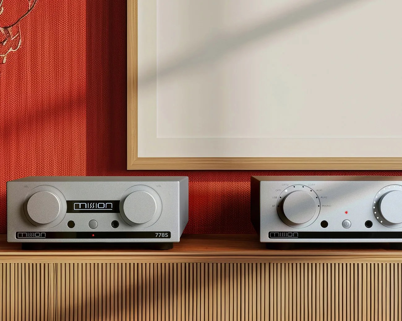 Two silver Mission audio components with large knobs on a wooden cabinet against a red textured wall and a framed artwork.