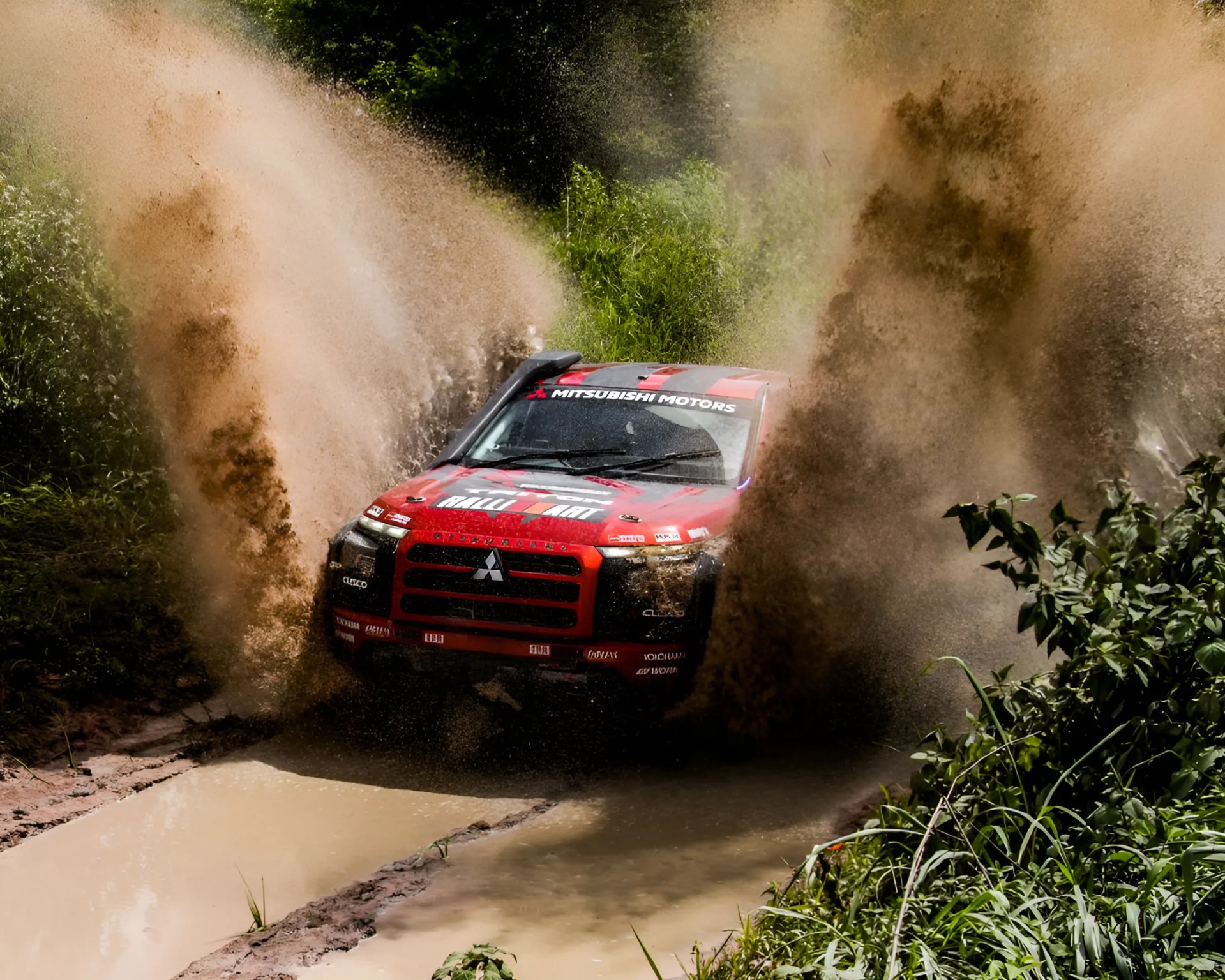 Red Mitsubishi rally car splashing mud and water while driving through a muddy forest trail.
