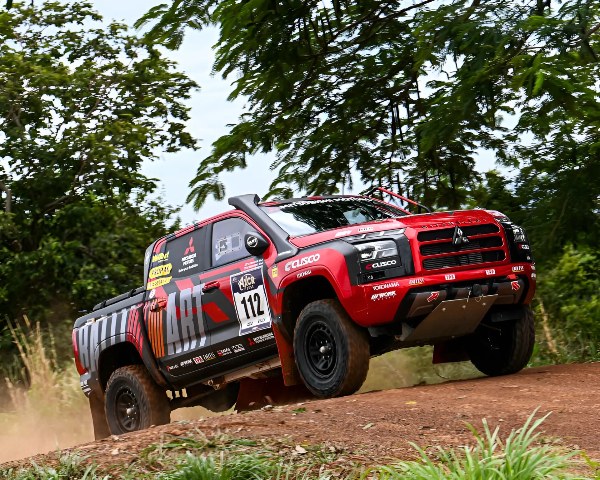 Red and black Mitsubishi rally truck with racing decals driving on a dirt road surrounded by trees.