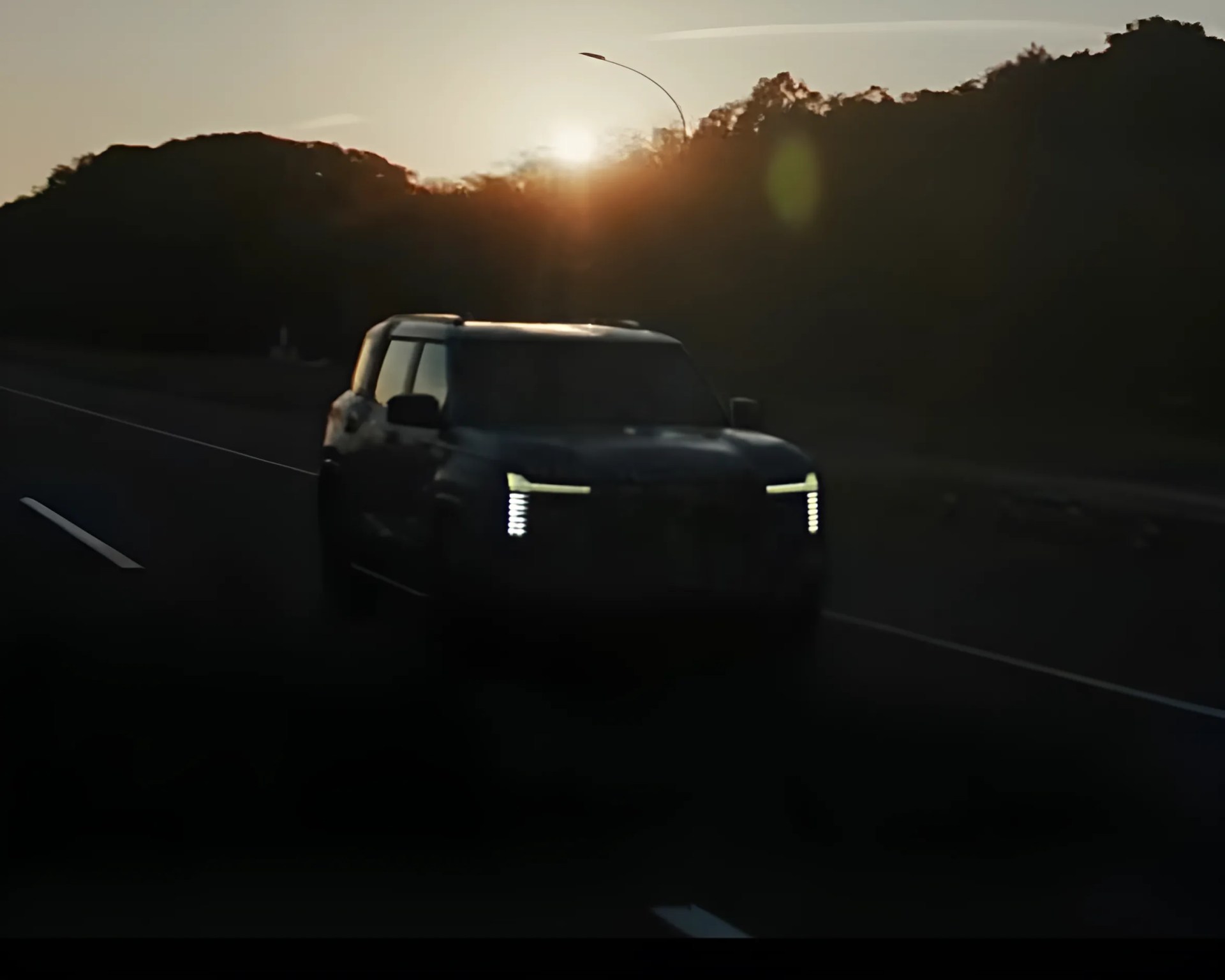 Dark SUV with vertical LED headlights driving on a road at sunset with hills in the background.