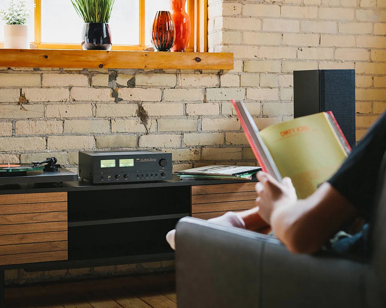 Person sitting on a gray chair reading a book near a wooden cabinet with audio equipment and a brick wall background.