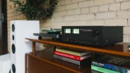 Black NAD stereophonic amplifier with knobs and meters on a wooden shelf next to stacked books and a white speaker.