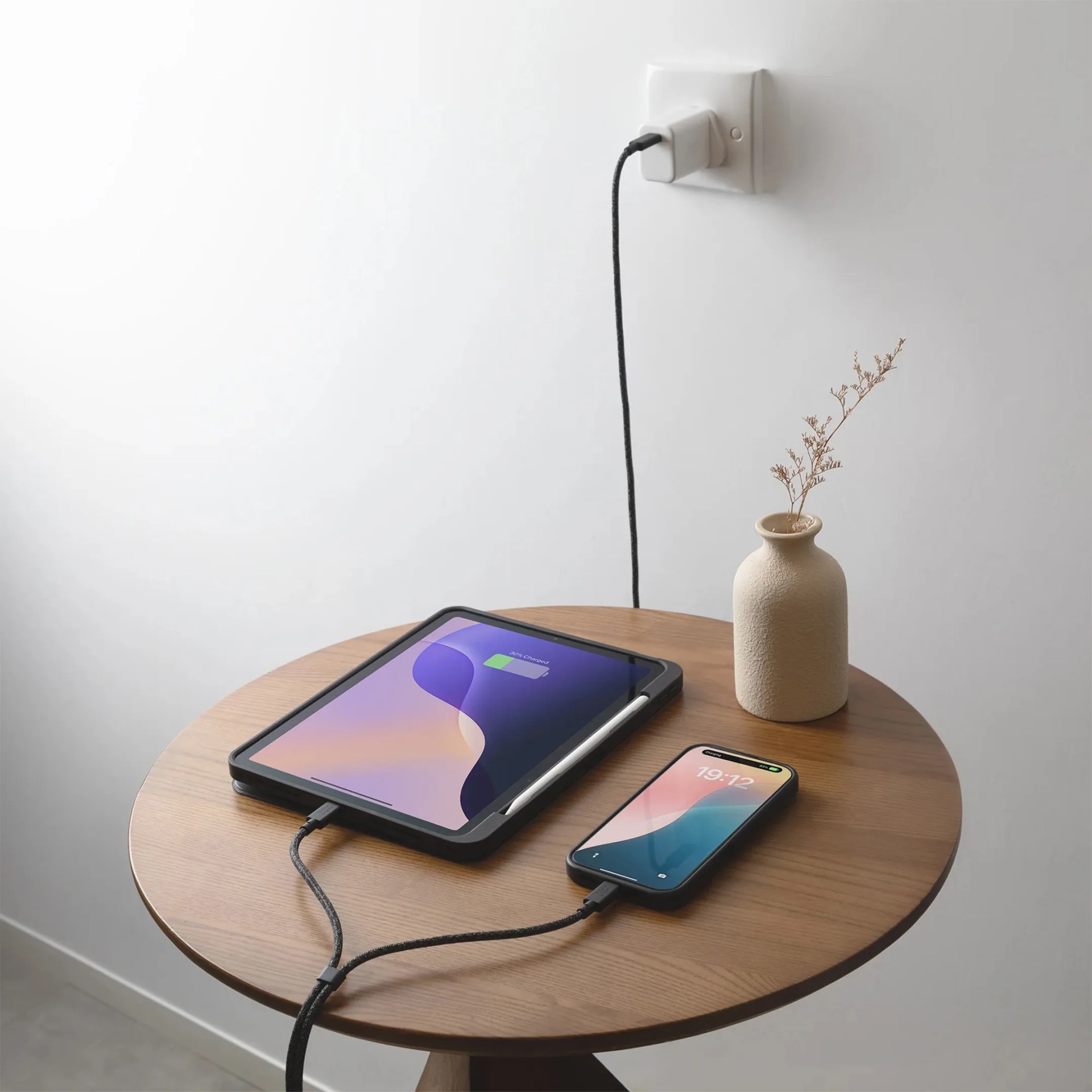 Wooden round table with a tablet and smartphone charging via a black braided splitter cable plugged into a wall outlet, next to a beige vase with dried flowers.