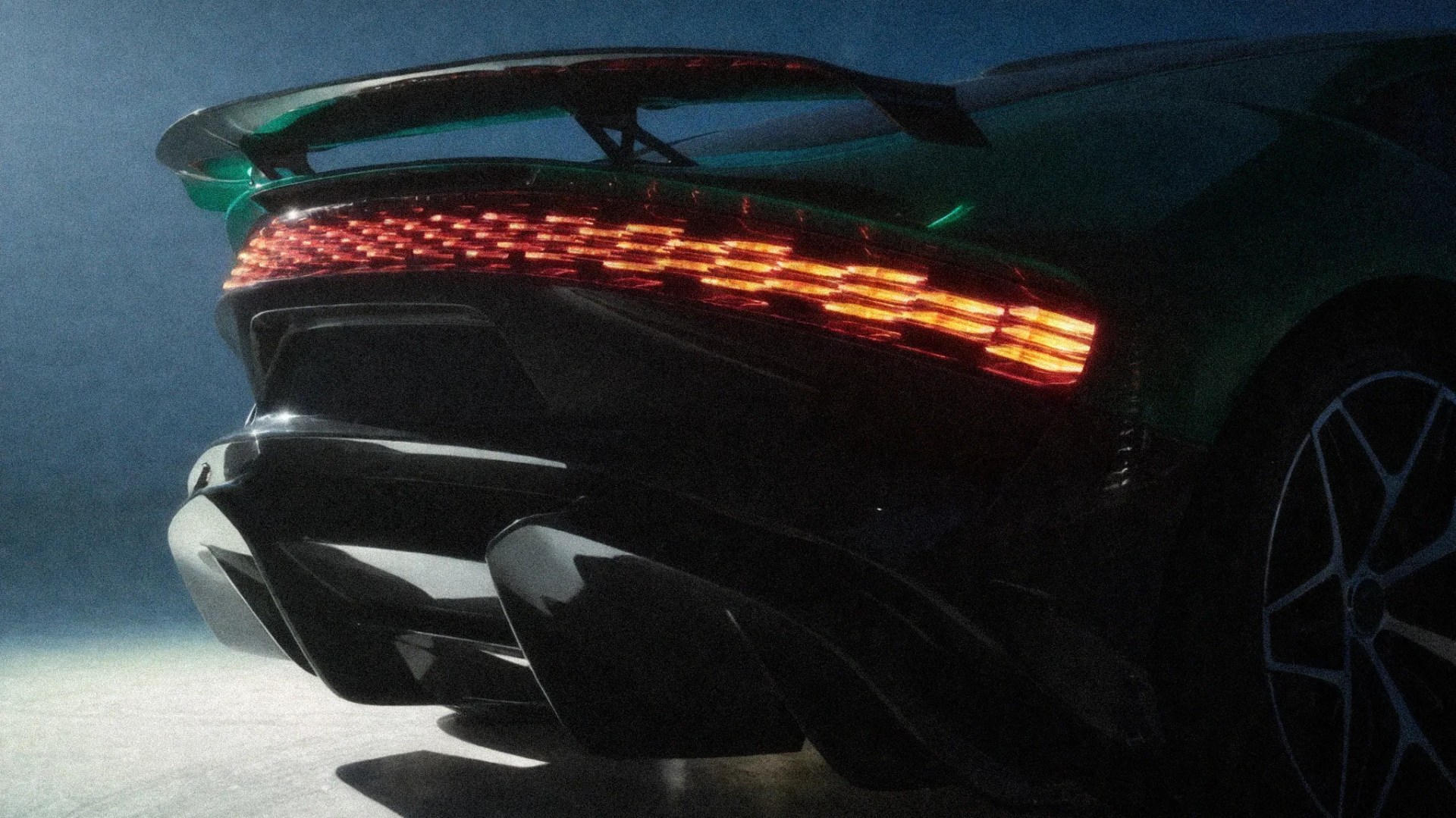 Rear view of a dark green sports car with illuminated red taillights, a large rear wing, and aggressive diffuser.