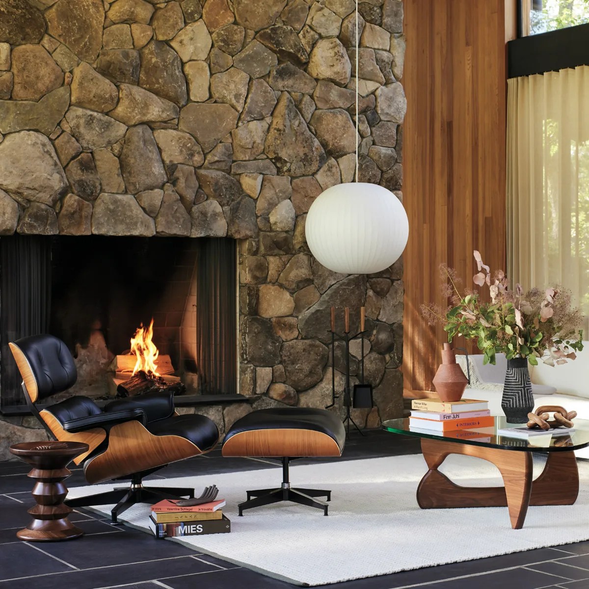 Mid-century modern living room with black leather and wood lounge chair, matching ottoman, stone fireplace, and glass coffee table with wooden base.