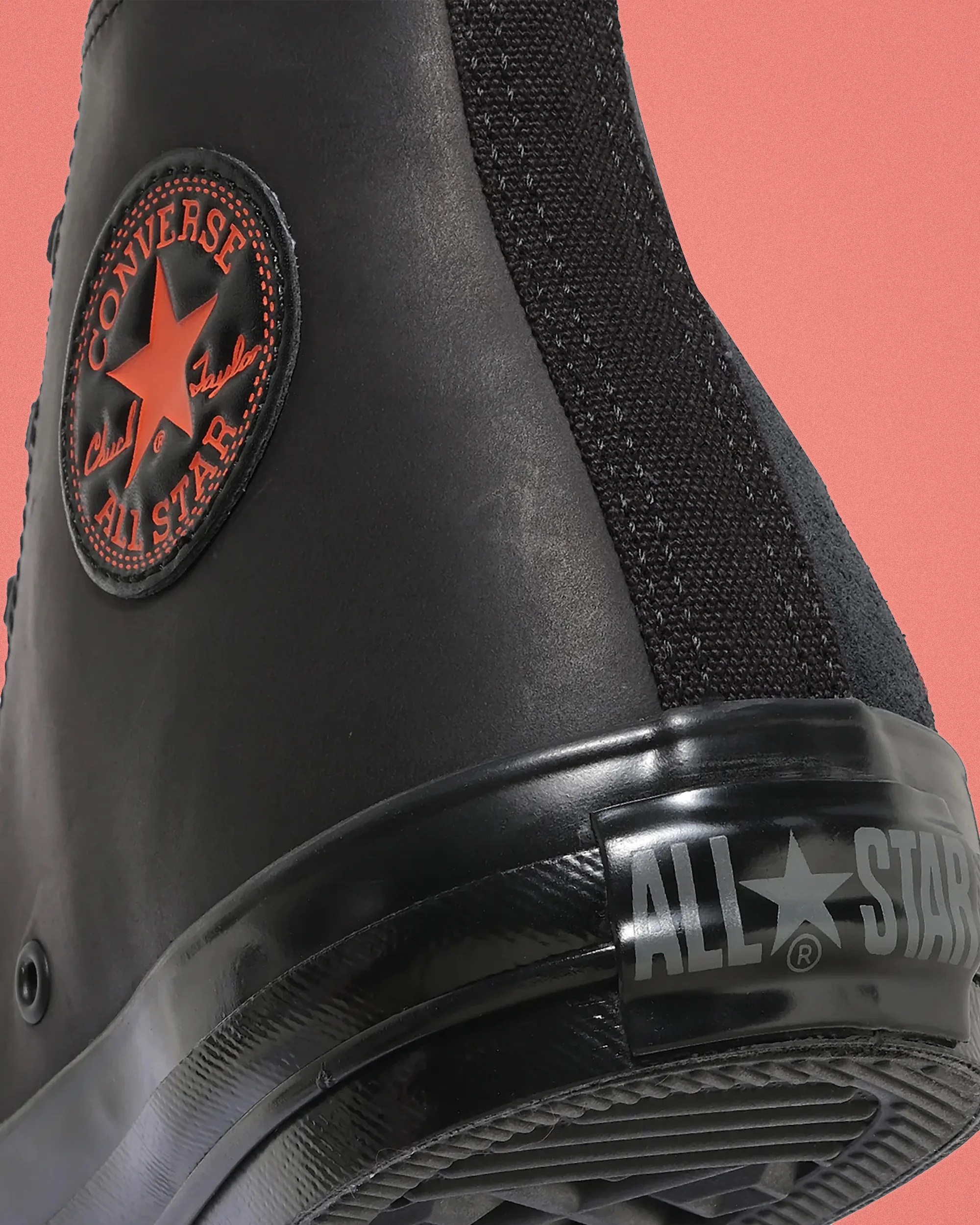 Close-up of the black Converse All Star sneaker heel with red logo patch and black rubber sole against a coral background.