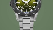 Close-up of a stainless steel watch with a green dial, white markers, and a black rotating bezel showing compass directions.