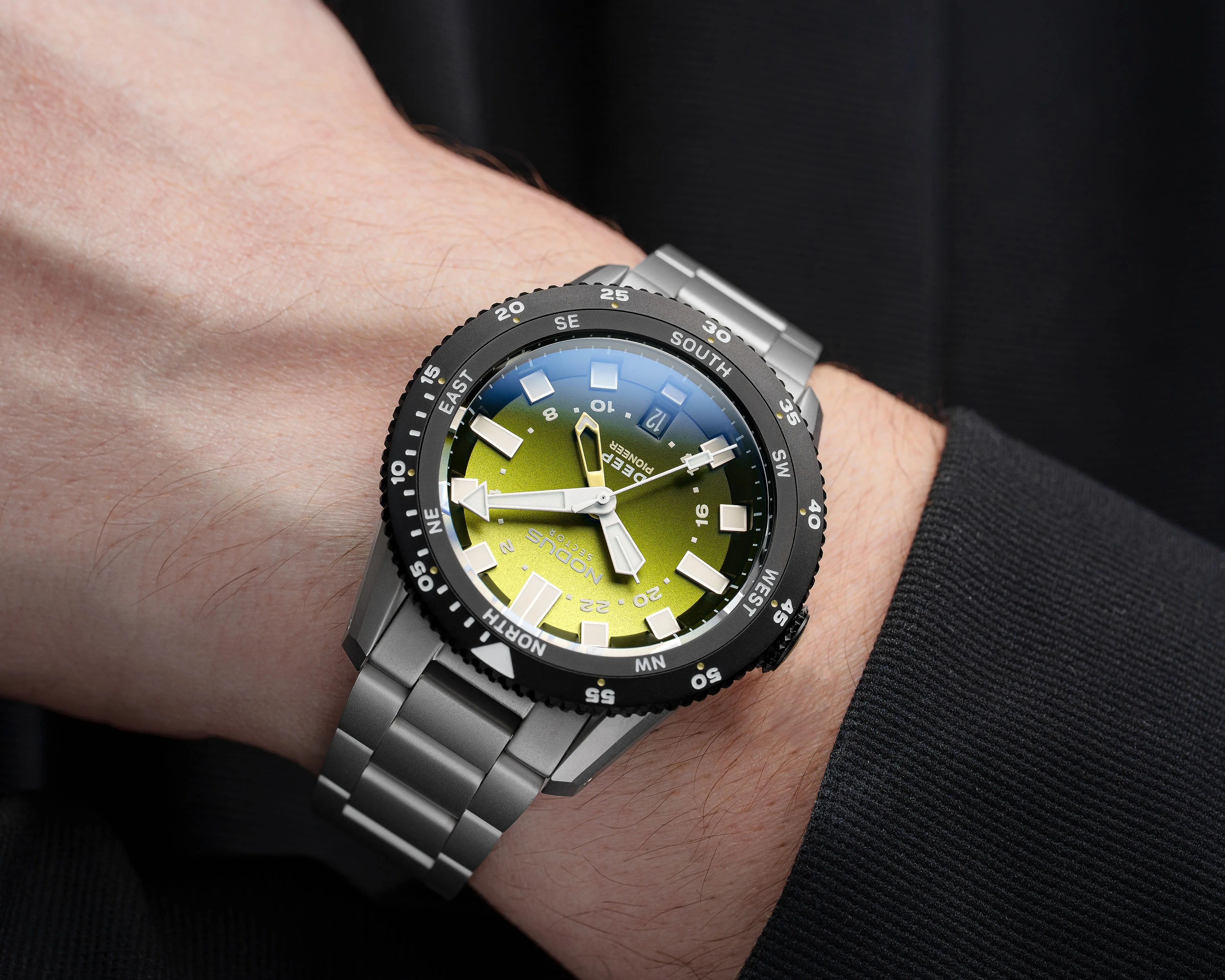 Silver metal wristwatch with a green gradient dial and black bezel showing compass directions, worn on a wrist.