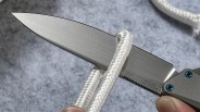 Close-up of a folding knife blade cutting a white braided rope against a gray background.